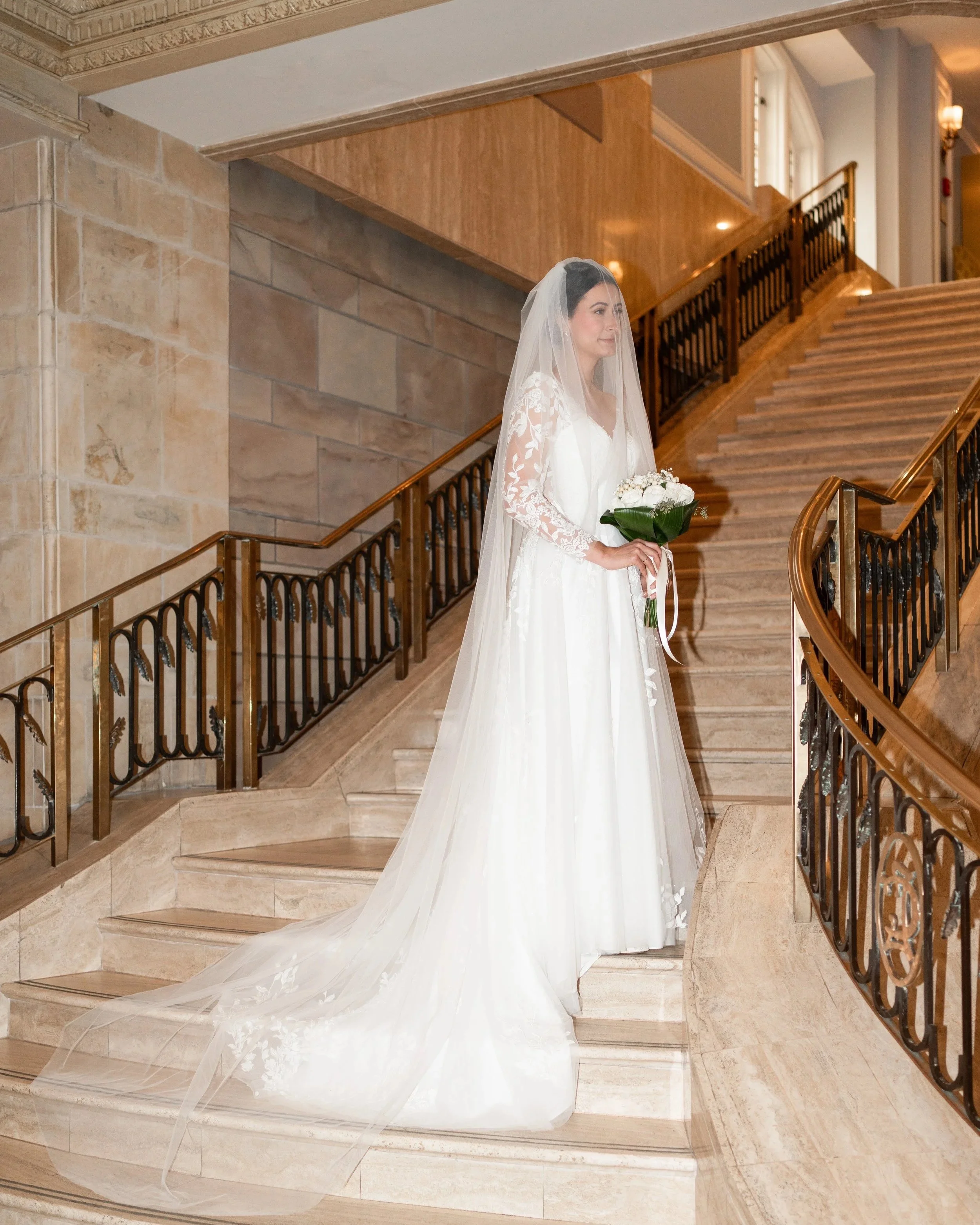 A bride in a white wedding dress with a long veil, holding a bouquet of white flowers, standing on a staircase inside a building with classic interior decor.