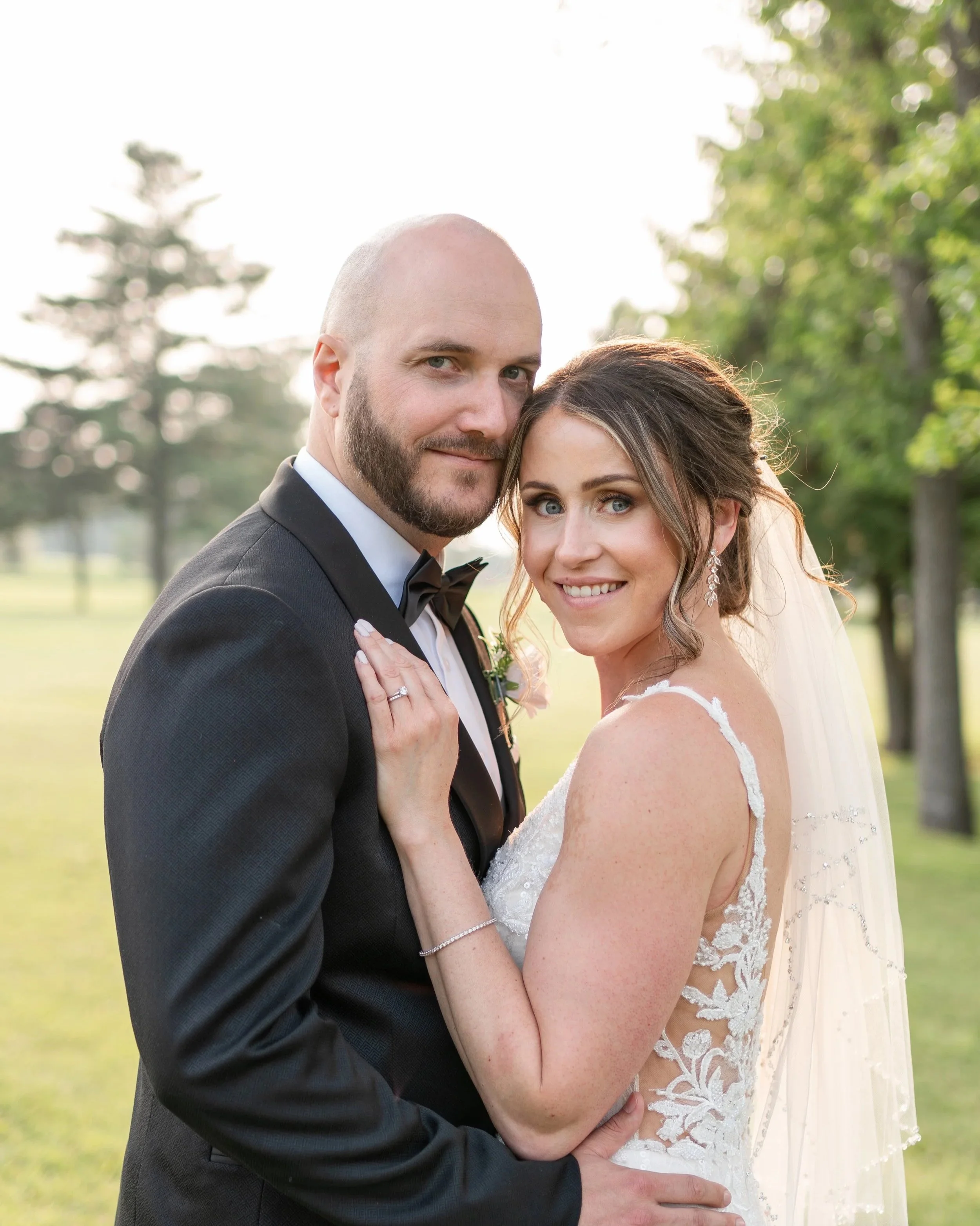 A bride and groom standing outdoors on a sunny day, smiling and embracing each other on their wedding day, with trees in the background.