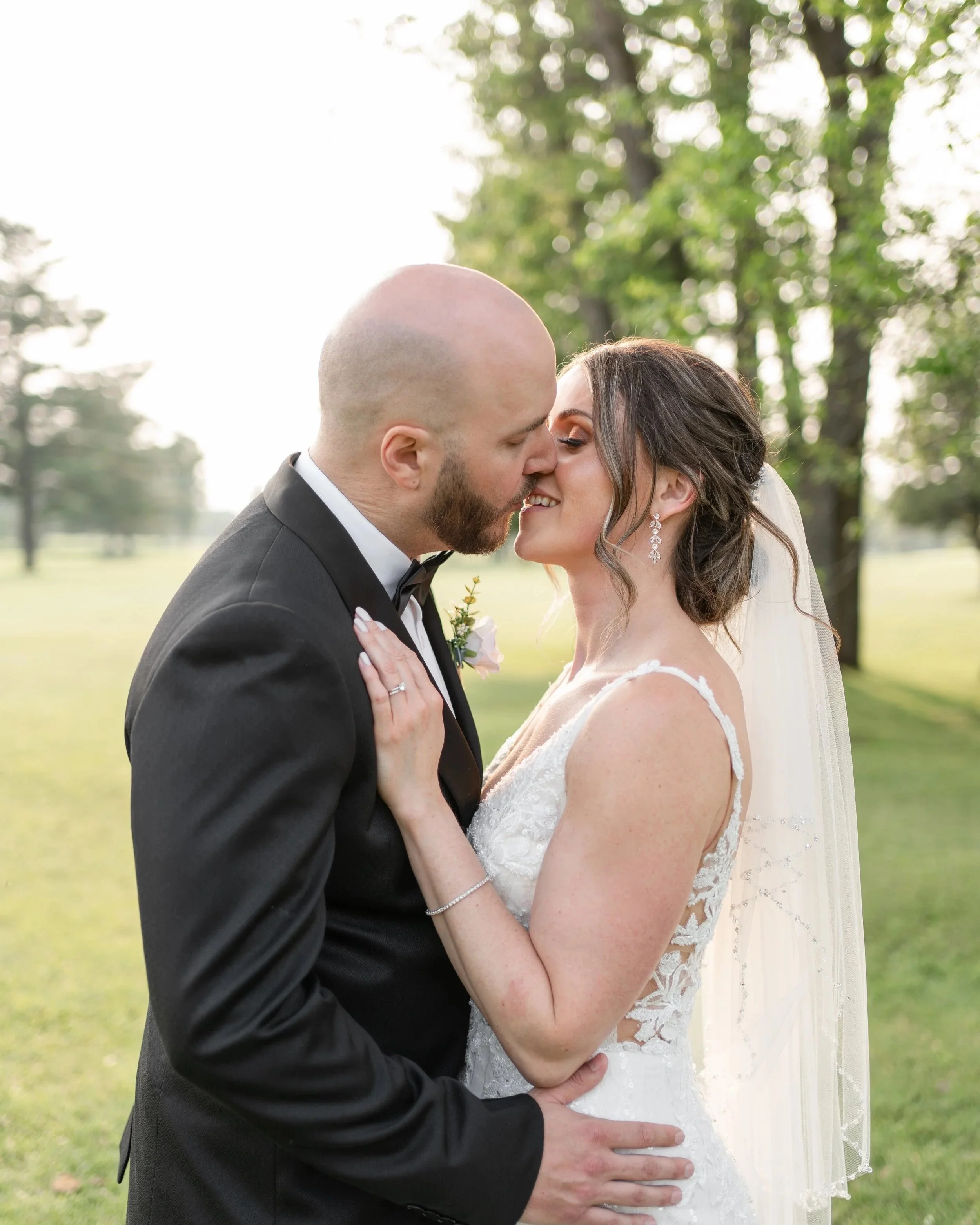 A newlywed couple is kissing outdoors in a park with green trees in the background.
