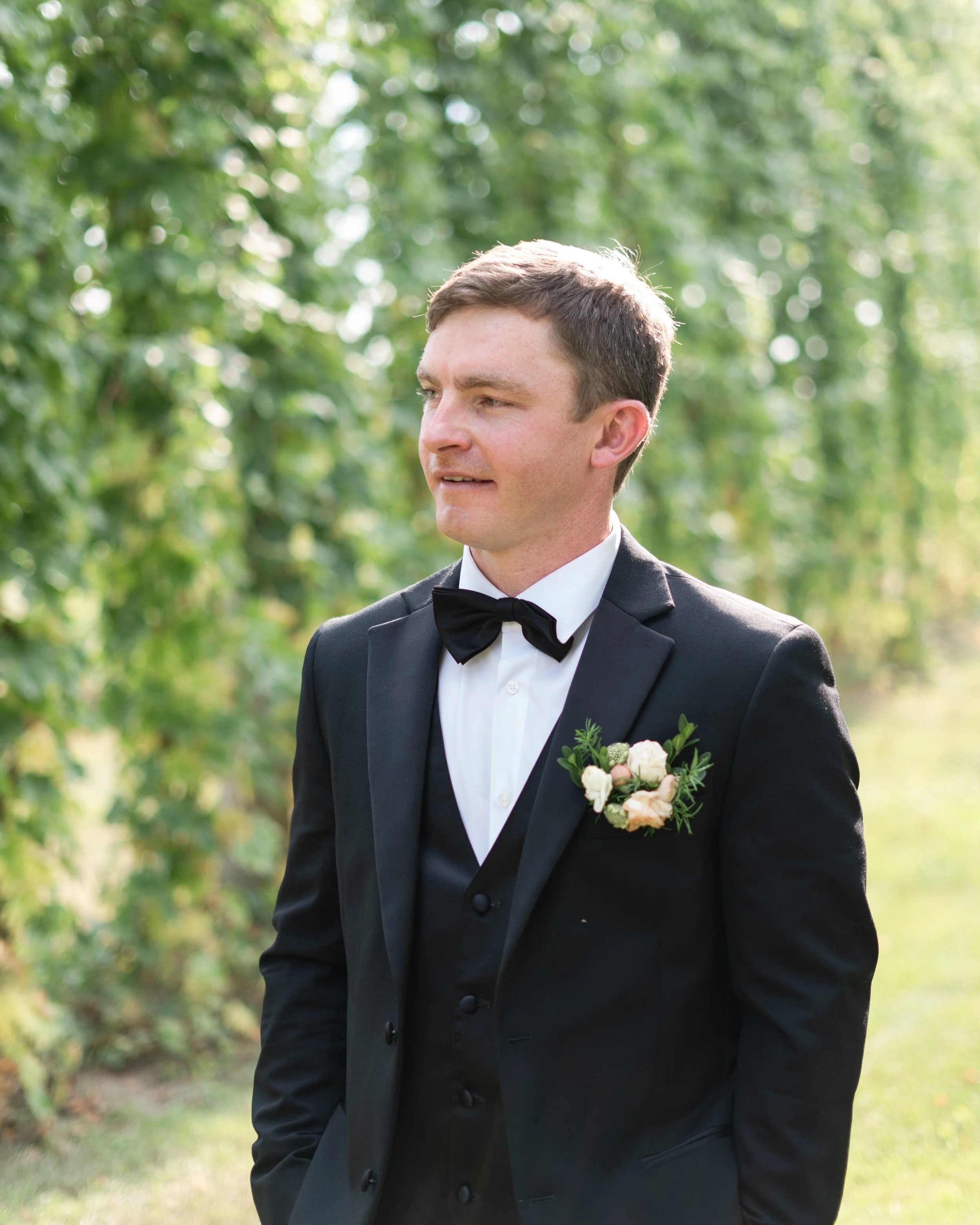 A groom in a black tuxedo with a boutonniere standing outdoors with greenery in the background. Stone Hedges Hops, Pembroke, ON