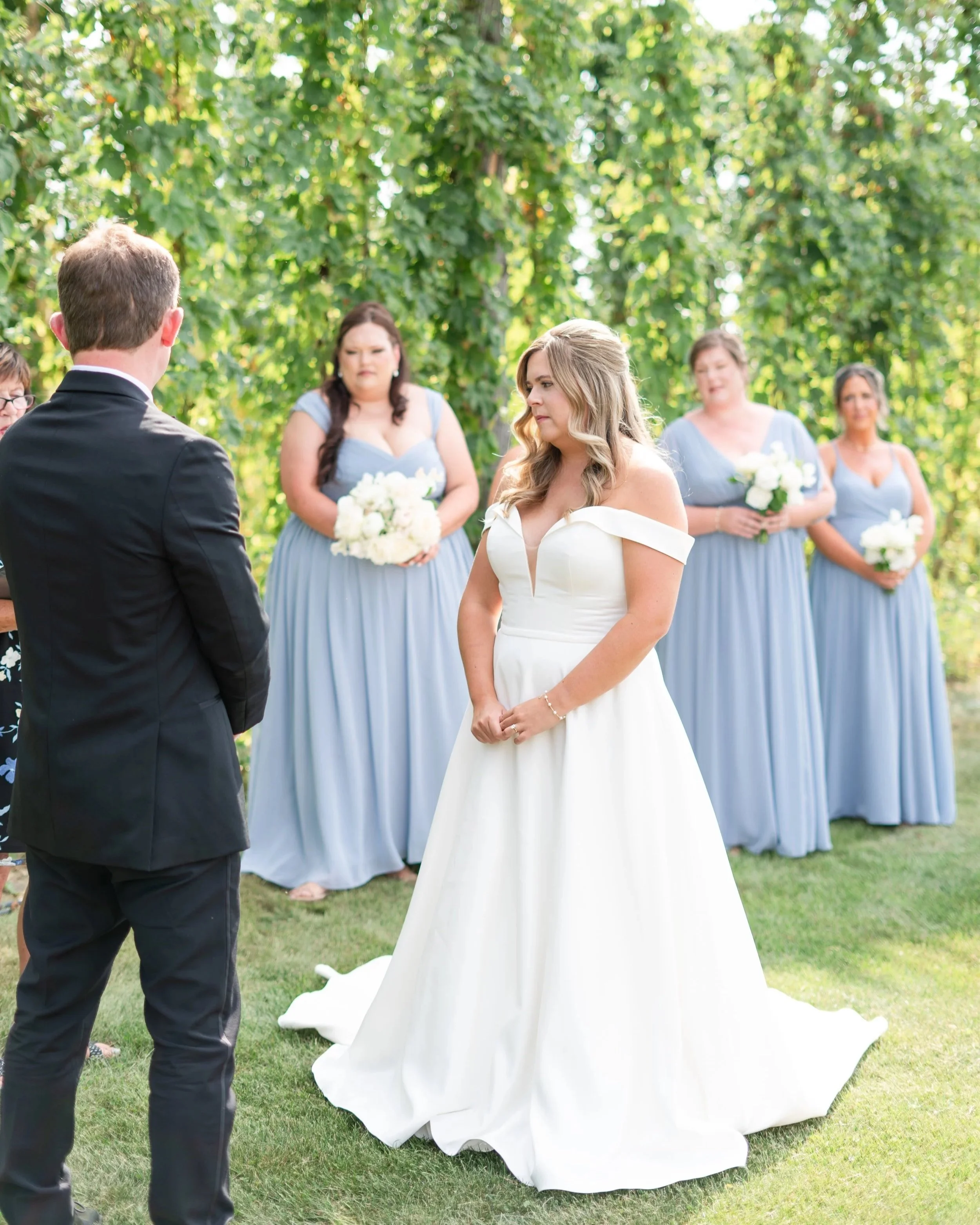 Bride and groom exchanging vows during an outdoor wedding ceremony, with bridesmaids holding white bouquets in the background. Stone Hedges Hops, Pembroke, ON