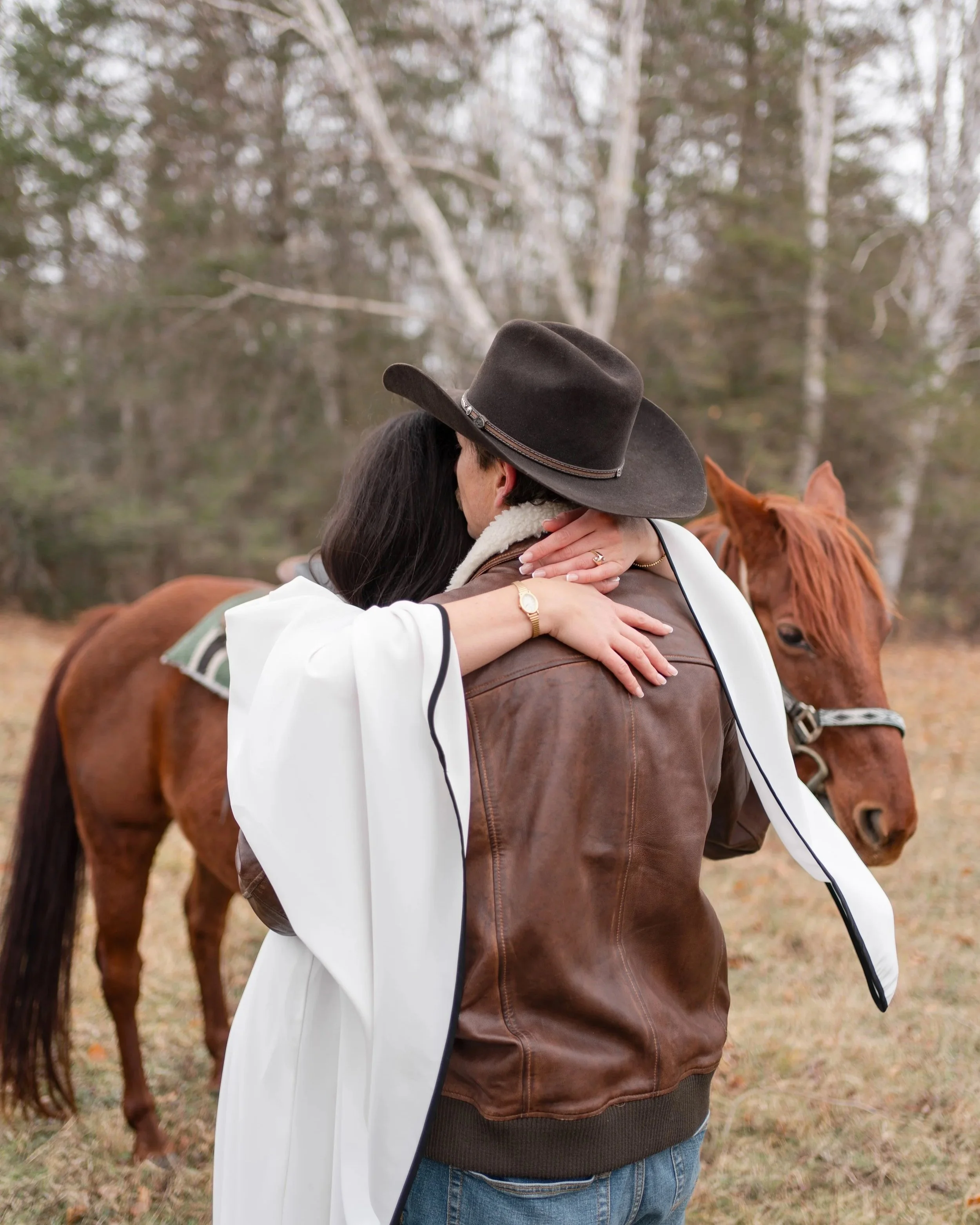A couple hugging outdoors with a brown horse nearby, surrounded by trees on a fall day.