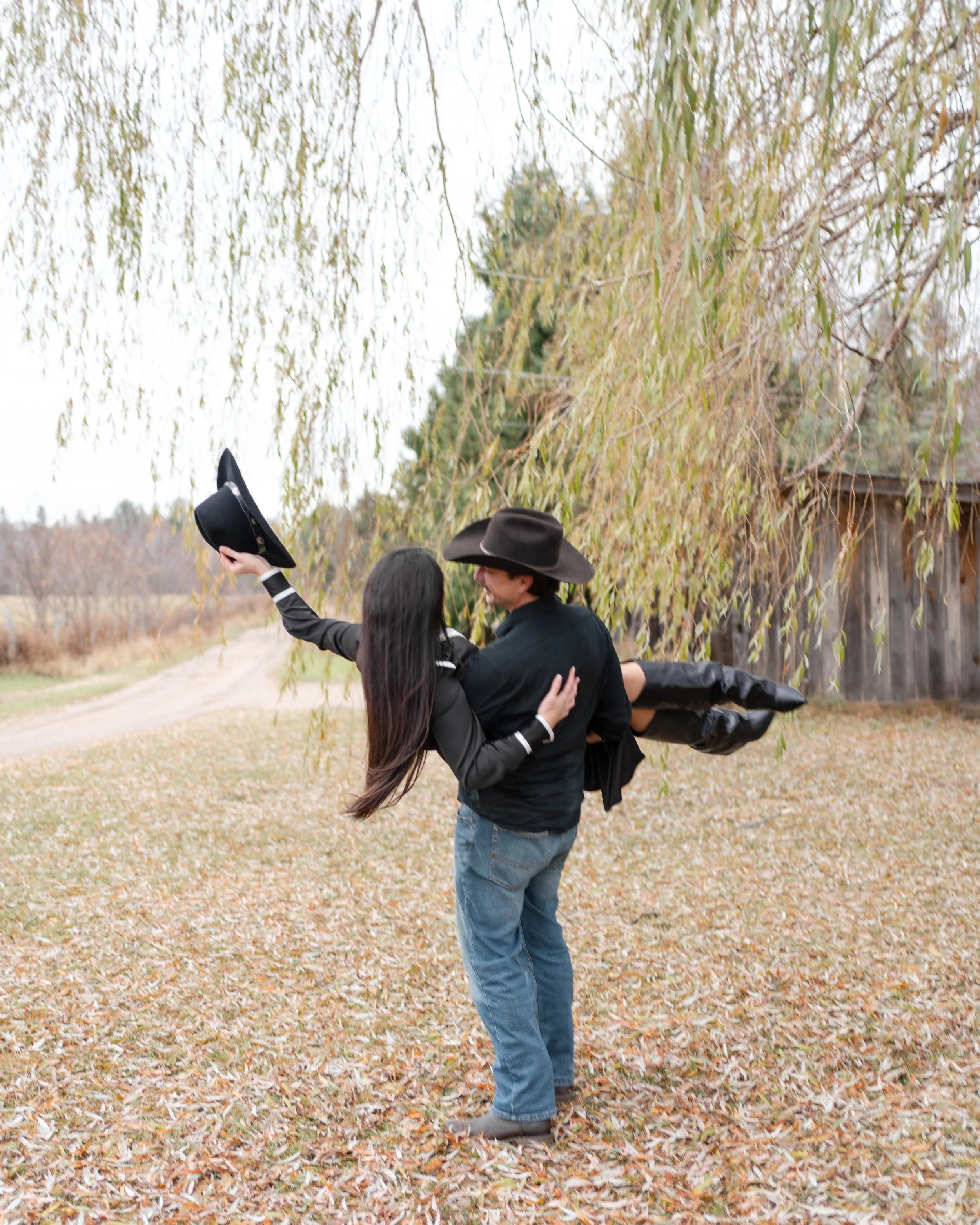 A man in a black cowboy hat is lifting a woman wearing a black jacket and black boots, holding her in his arms outdoors near trees with autumn leaves.
