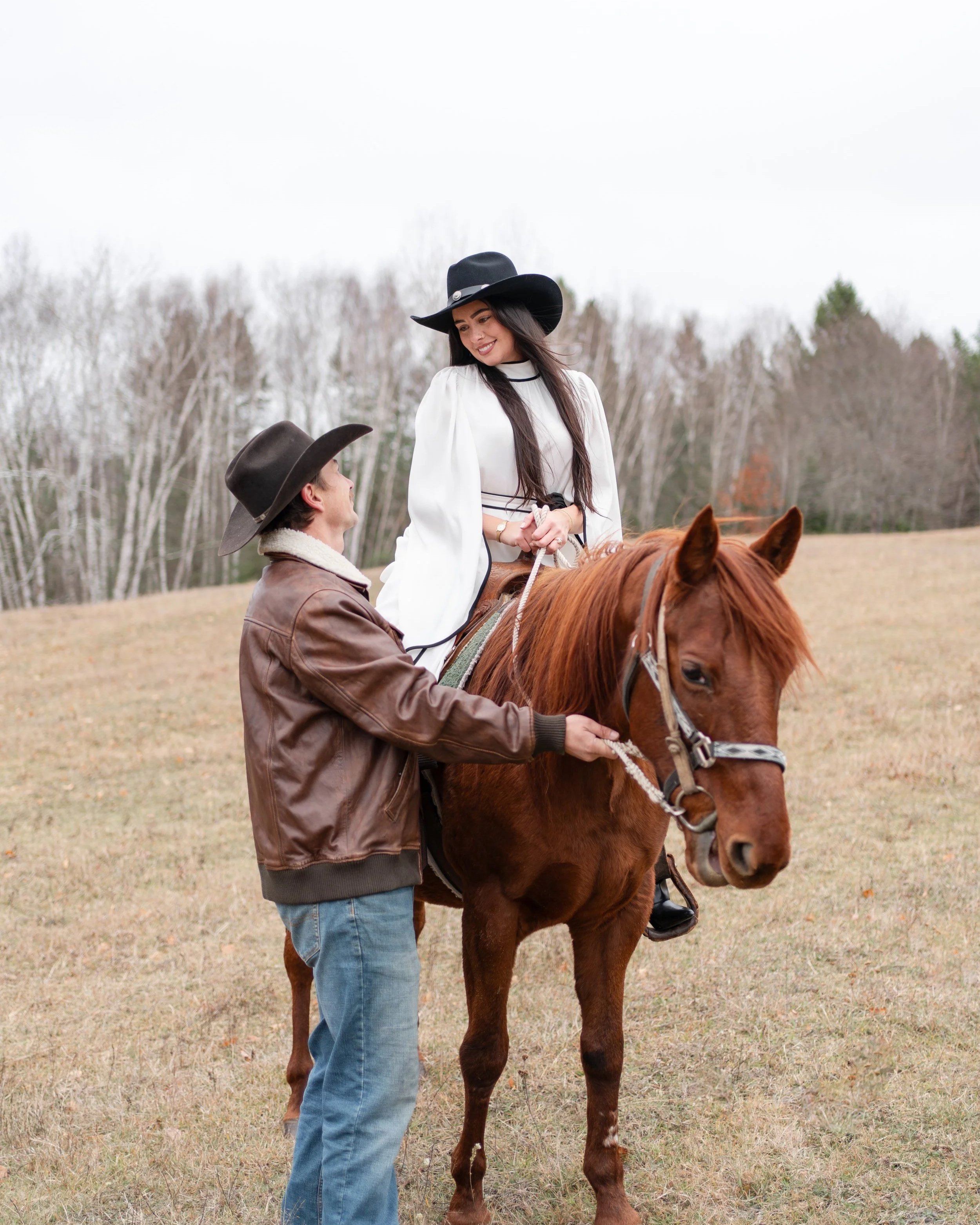 A woman sitting on a horse being assisted by a man in an open field with trees in the background.