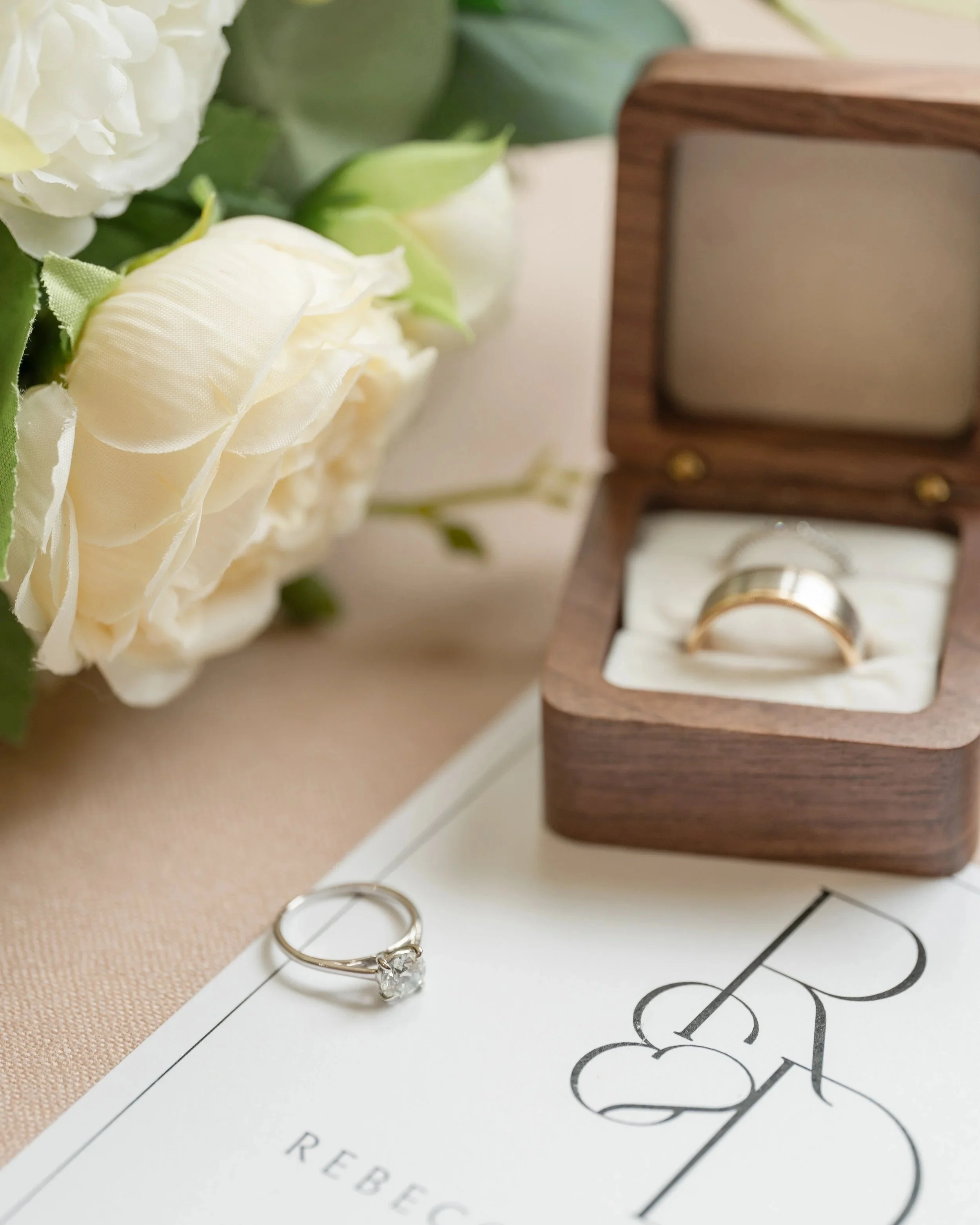Wedding ring in a wooden box, engagement ring with a diamond, bouquet of white roses, and wedding invitation on a beige surface.