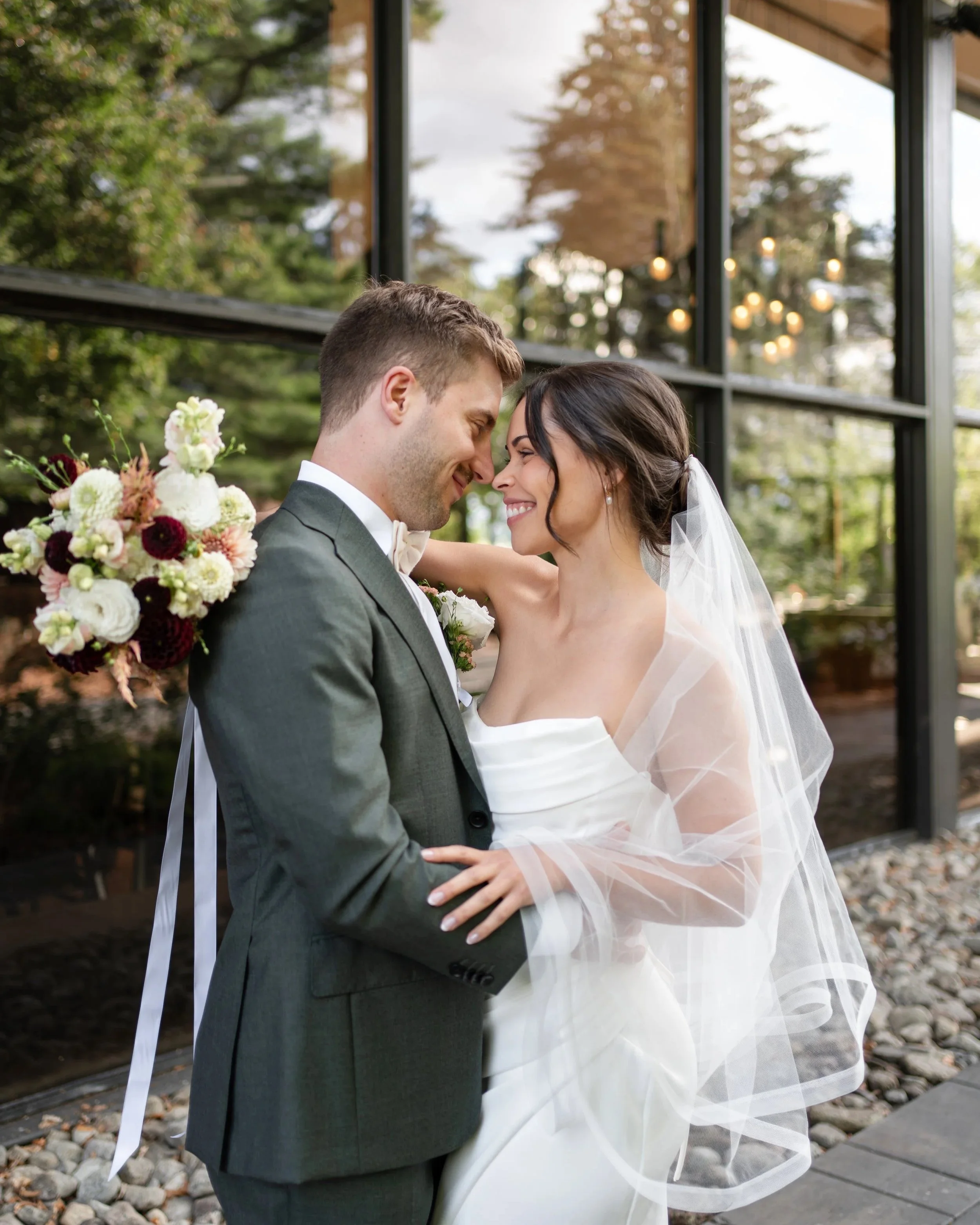 A bride and groom on their wedding day with their foreheads touching and smiling. The bride wears a strapless white wedding dress and veil, holding a bouquet of flowers. The groom wears a gray suit with a white shirt and bow tie, standing outside near a glass building with green trees in the background.