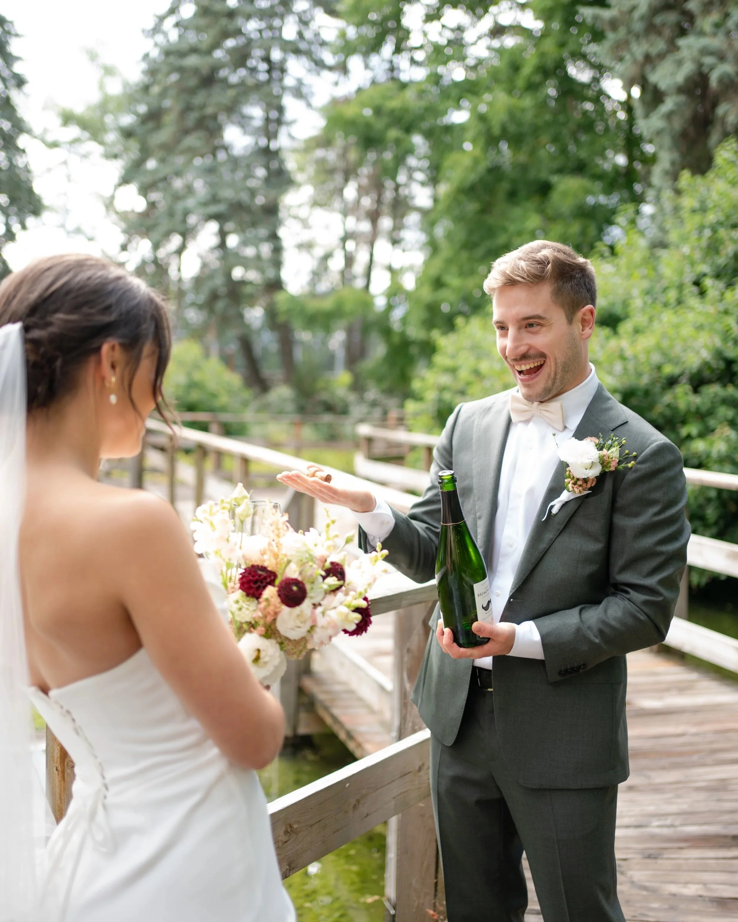 A groom in a gray suit and white bow tie is smiling and holding a champagne bottle. La Toundra, Montreal