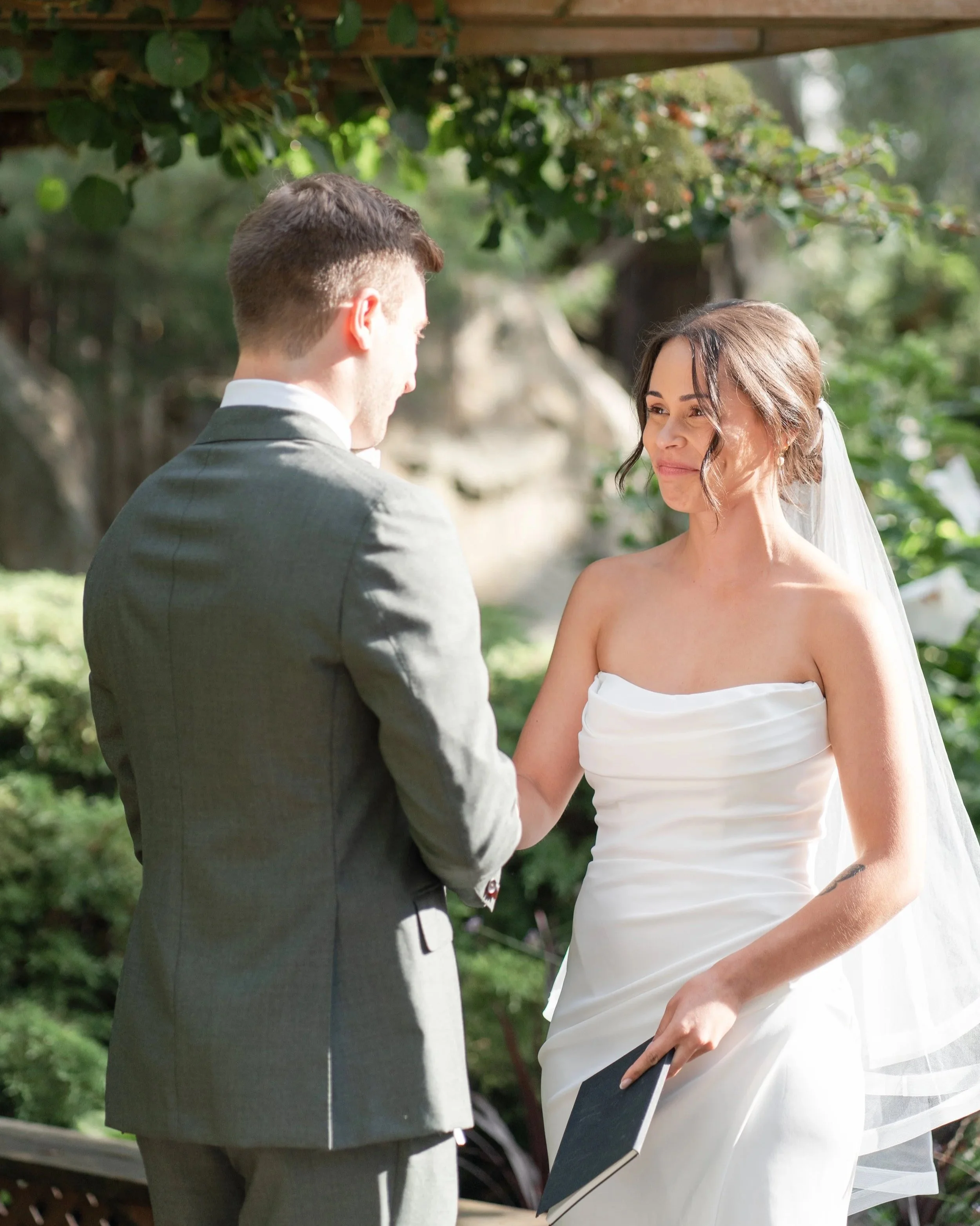A bride and groom during their wedding ceremony outdoors in the garden, holding hands and gazing at each other, with greenery and trees in the background. La toundra, Montreal