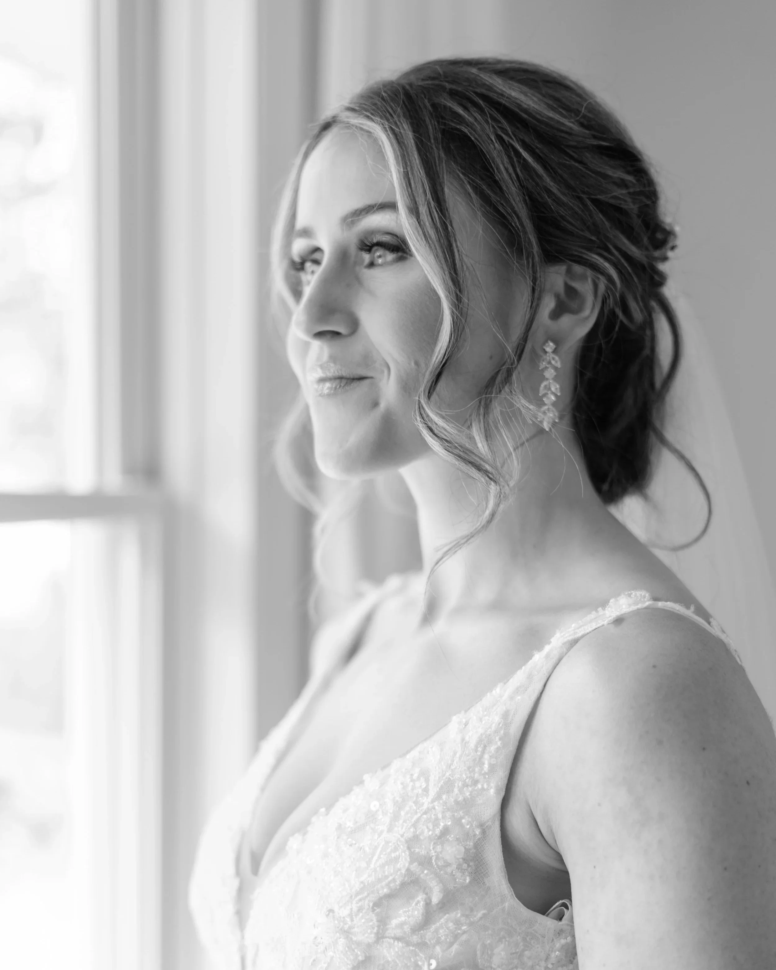 Black and white photo of a bride with a gentle smile, looking out a window, wearing earrings and a wedding dress.