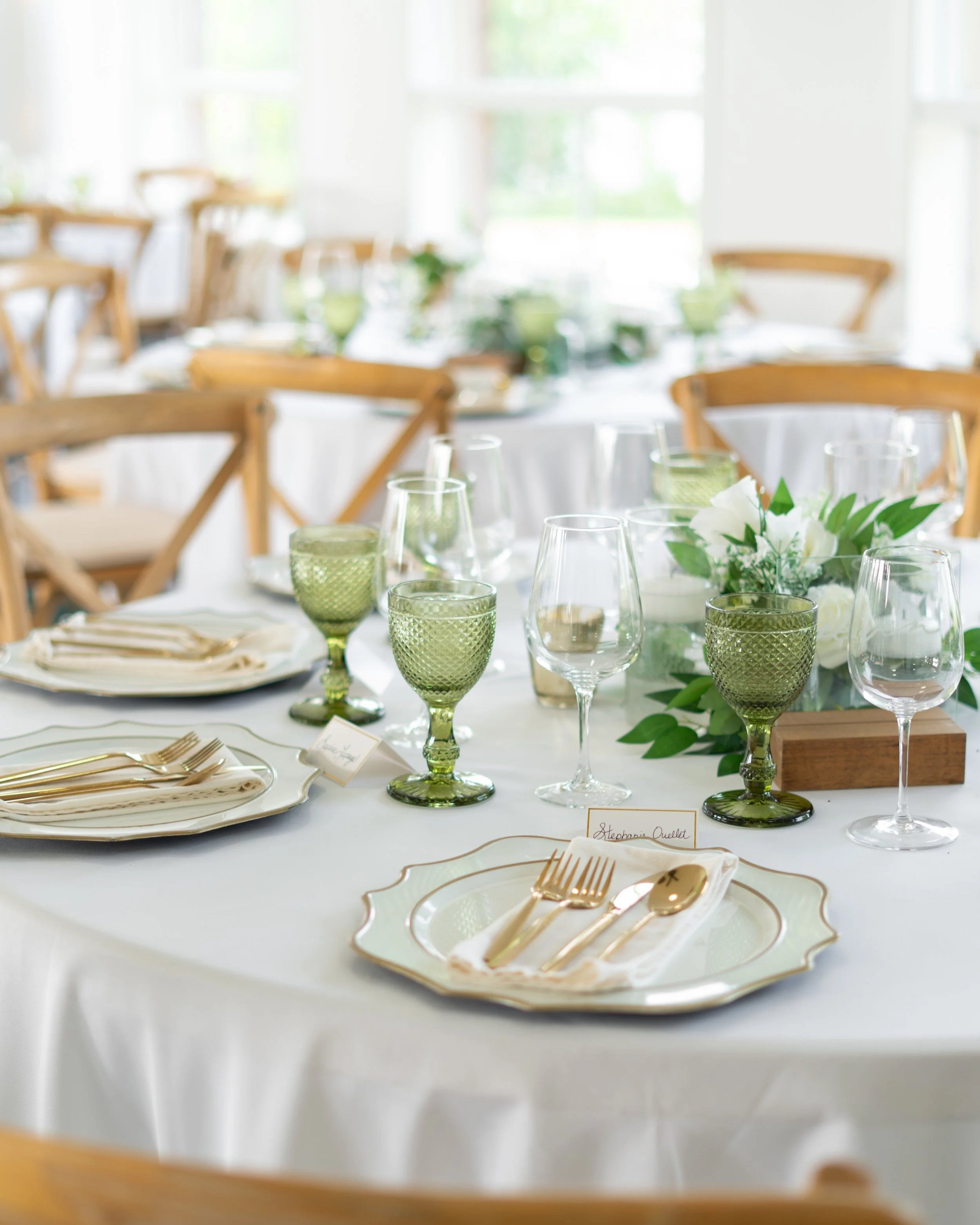 Elegant table setting with white tablecloth, gold cutlery, white plates, green and clear glassware, and a floral centerpiece with white flowers and green leaves in a bright room with wooden chairs.