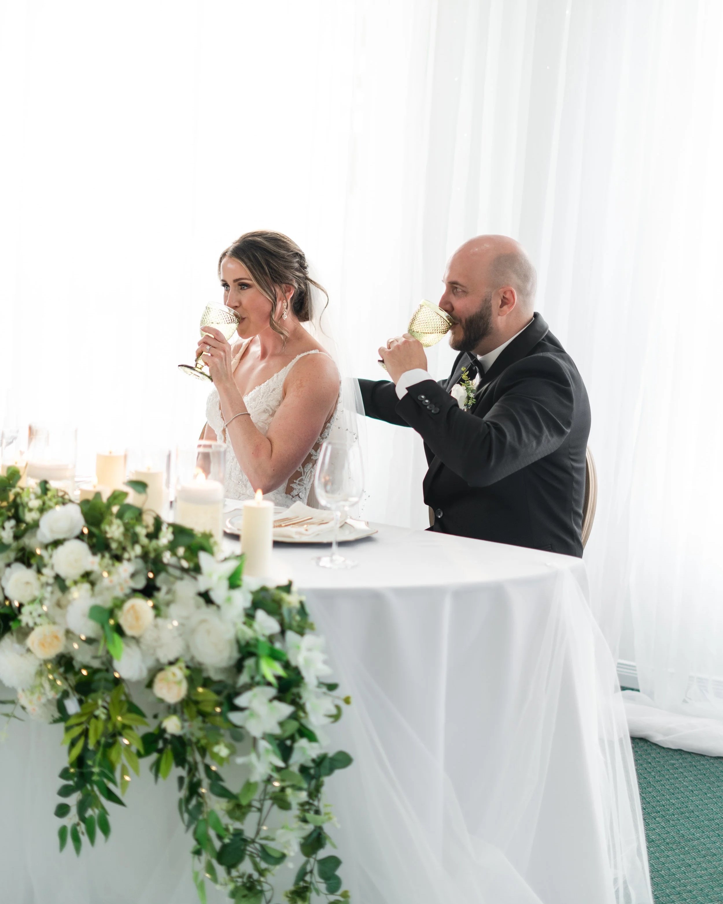 Bride and groom at wedding reception drinking from green-colored glasses, sitting at a decorated table with candles and flowers, white curtains in background.