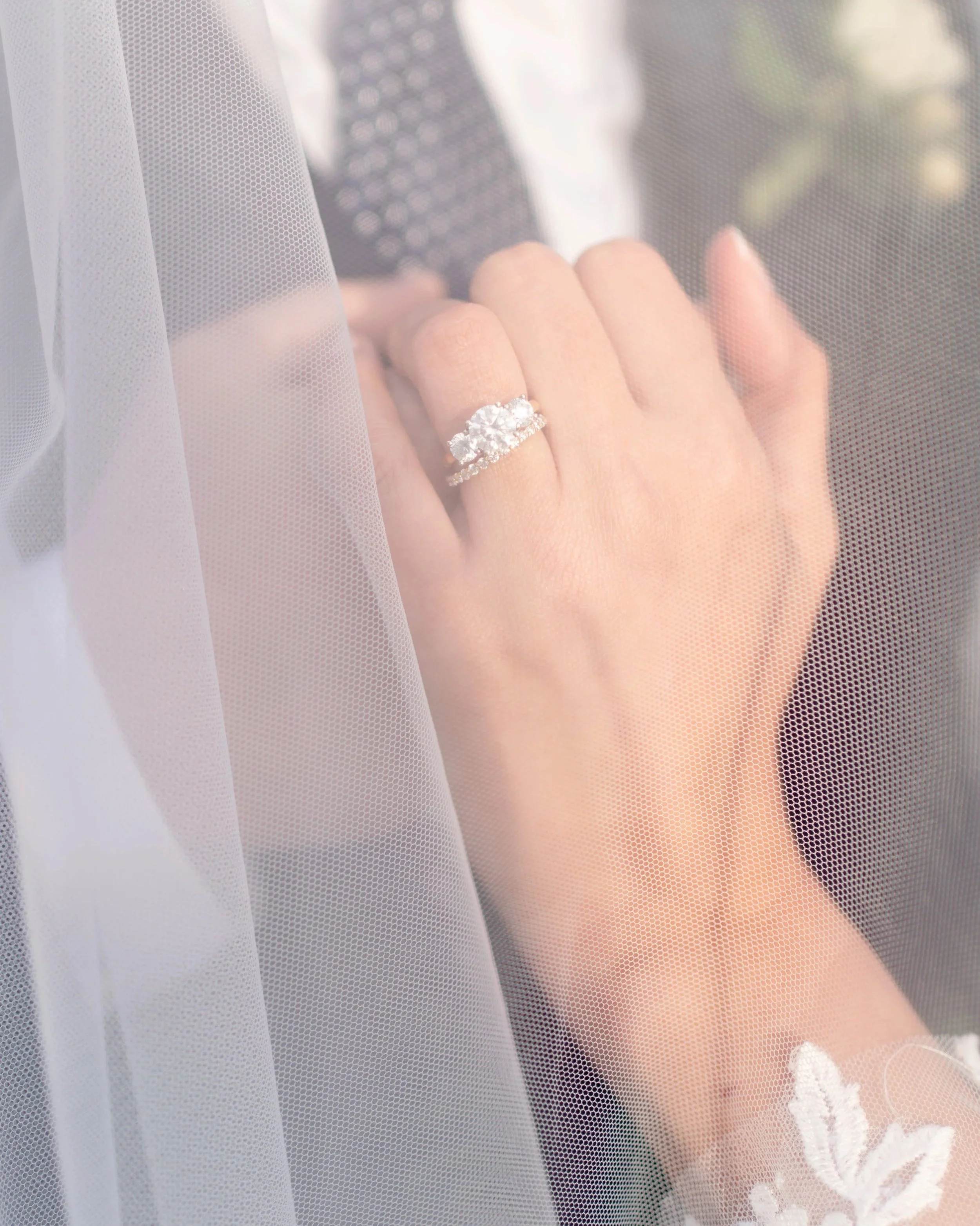 Close-up of a woman's hand with an elaborate diamond ring, seen through a sheer veil with lace detailing, suggesting a wedding or engagement setting.