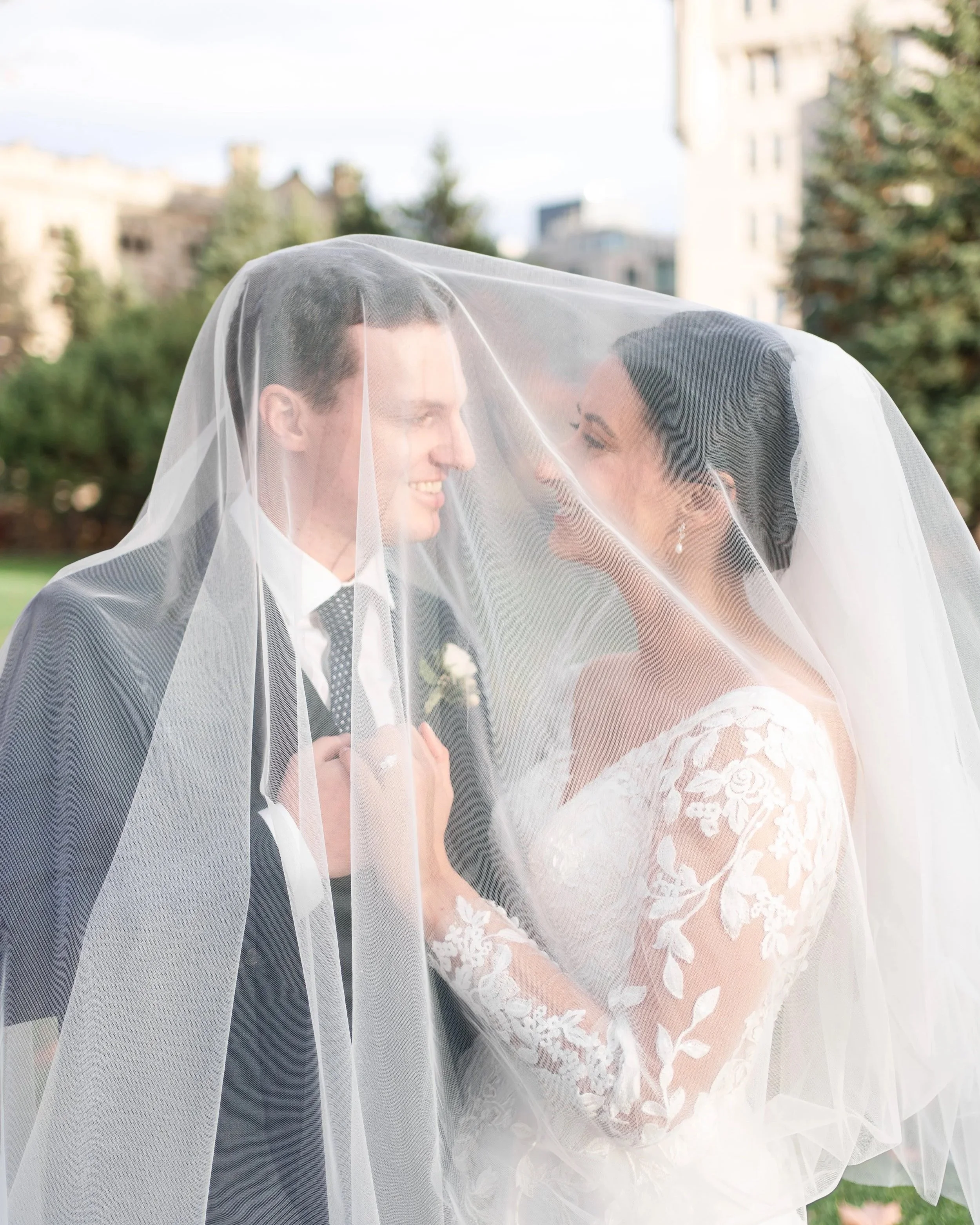 A newlywed couple smiling and touching under a sheer wedding veil outdoors with a cityscape and greenery in the background.