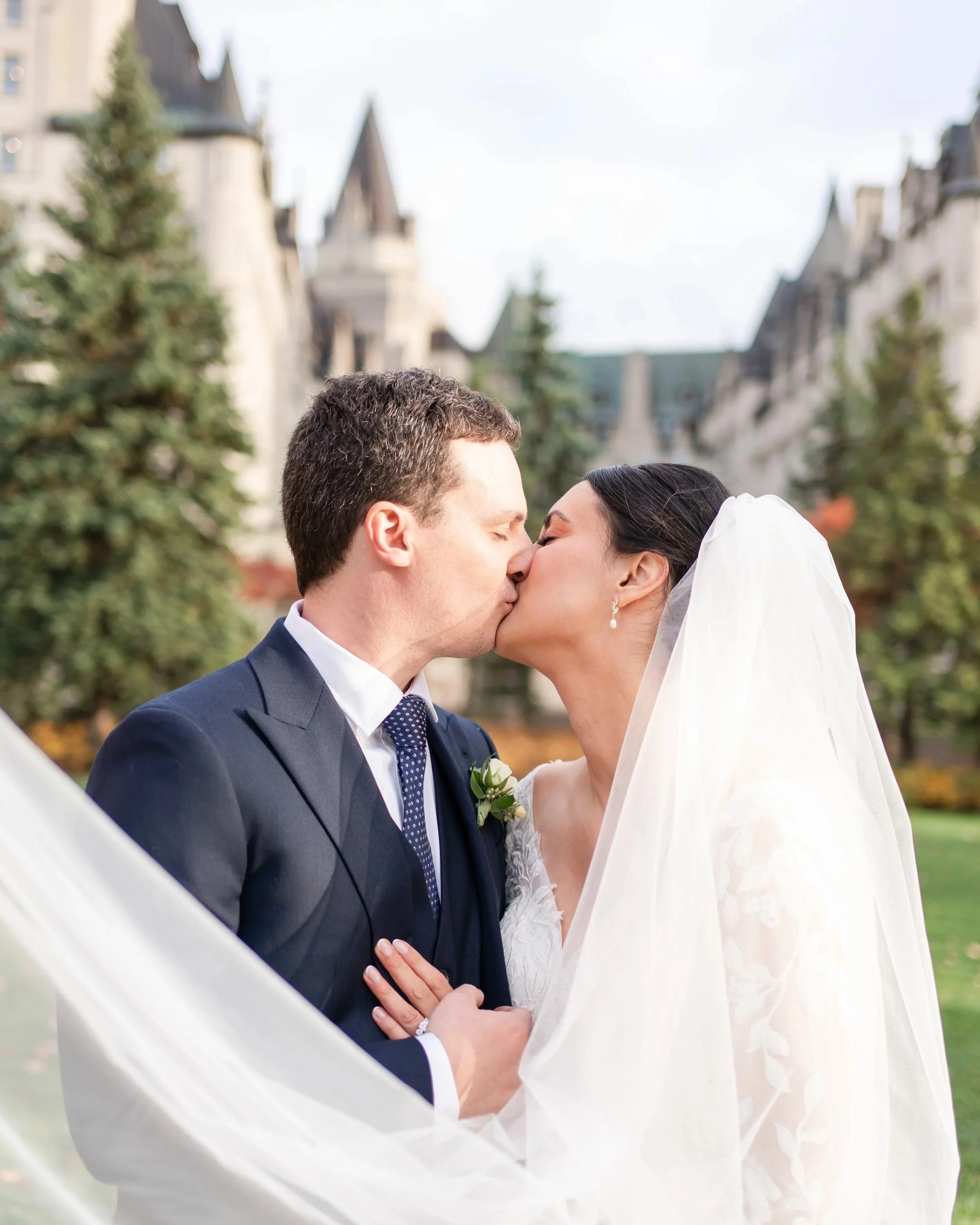 A bride and groom kissing outdoors with a castle-like building and trees in the background.