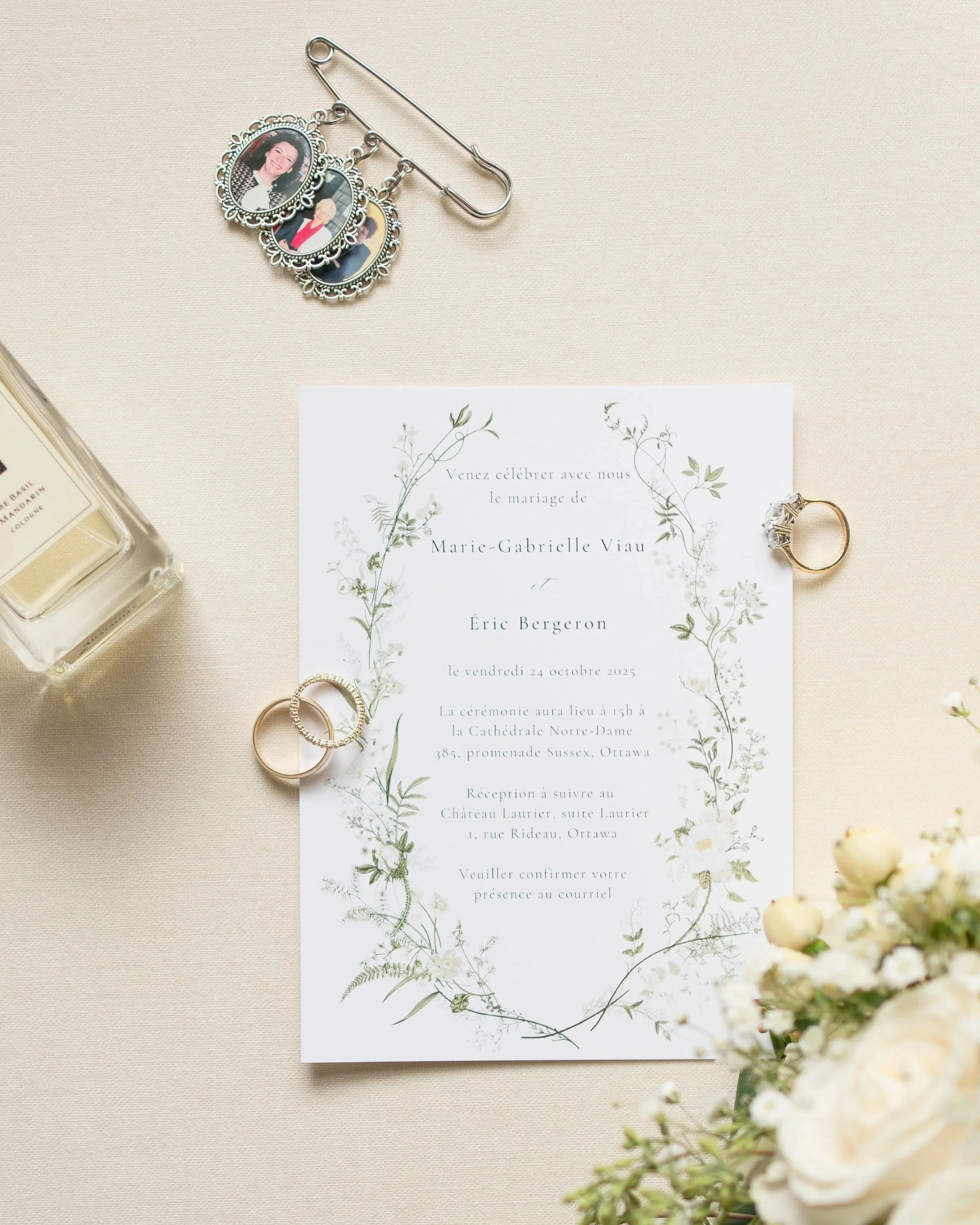 Wedding invitation with rings, a photo charm, flowers, perfume, and a safety pin with photo charms on a beige surface.