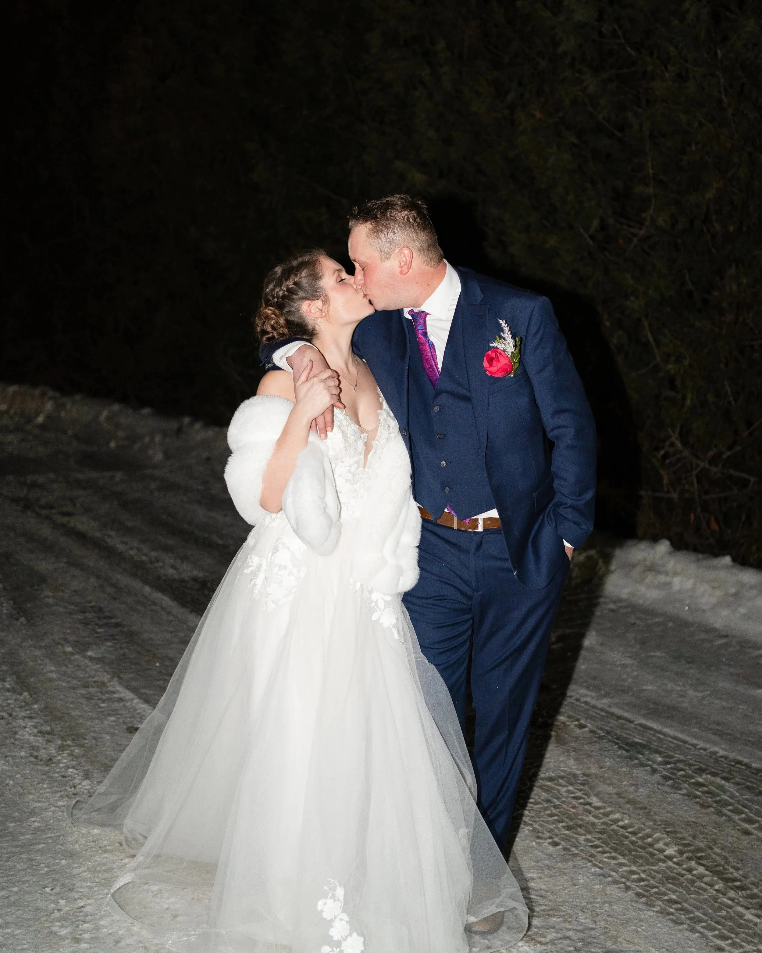 A bride and groom sharing a kiss outdoors at night, standing on a snow-covered road with dark trees in the background. Pembroke, ON