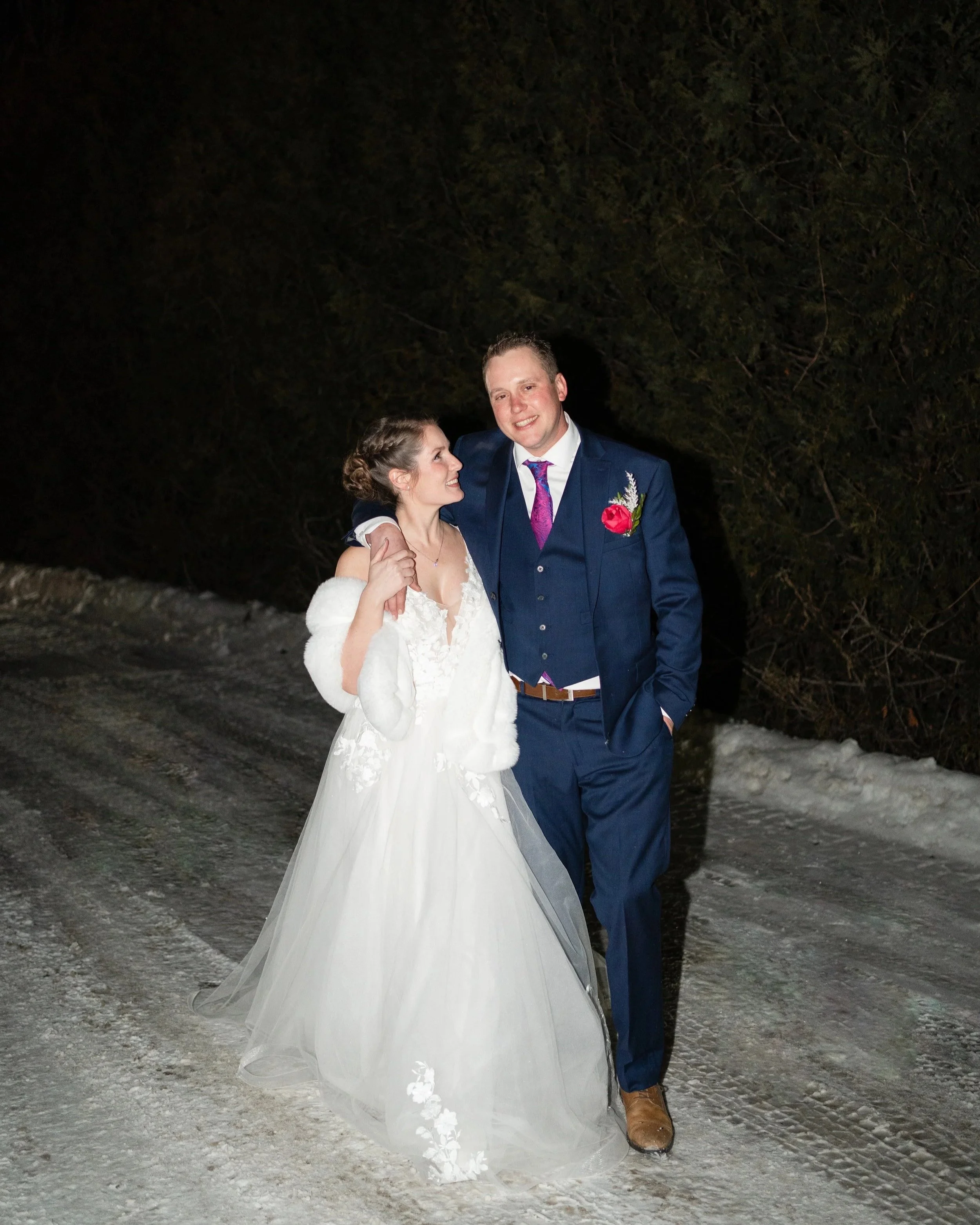 A bride and groom smiling and walking together on a snowy road at night, with trees in the background. Pembroke, Ontario.