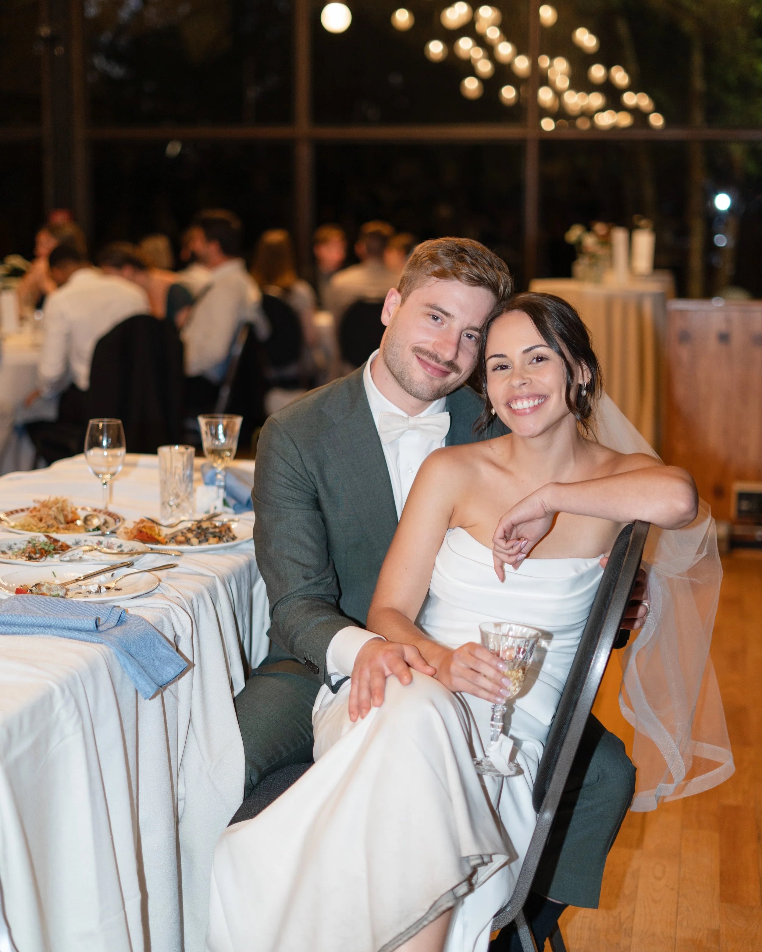 A newlywed couple at their wedding reception, sitting at a table with food and glasses, smiling, with guests in the background inside a decorated venue. La toundra, Montreal