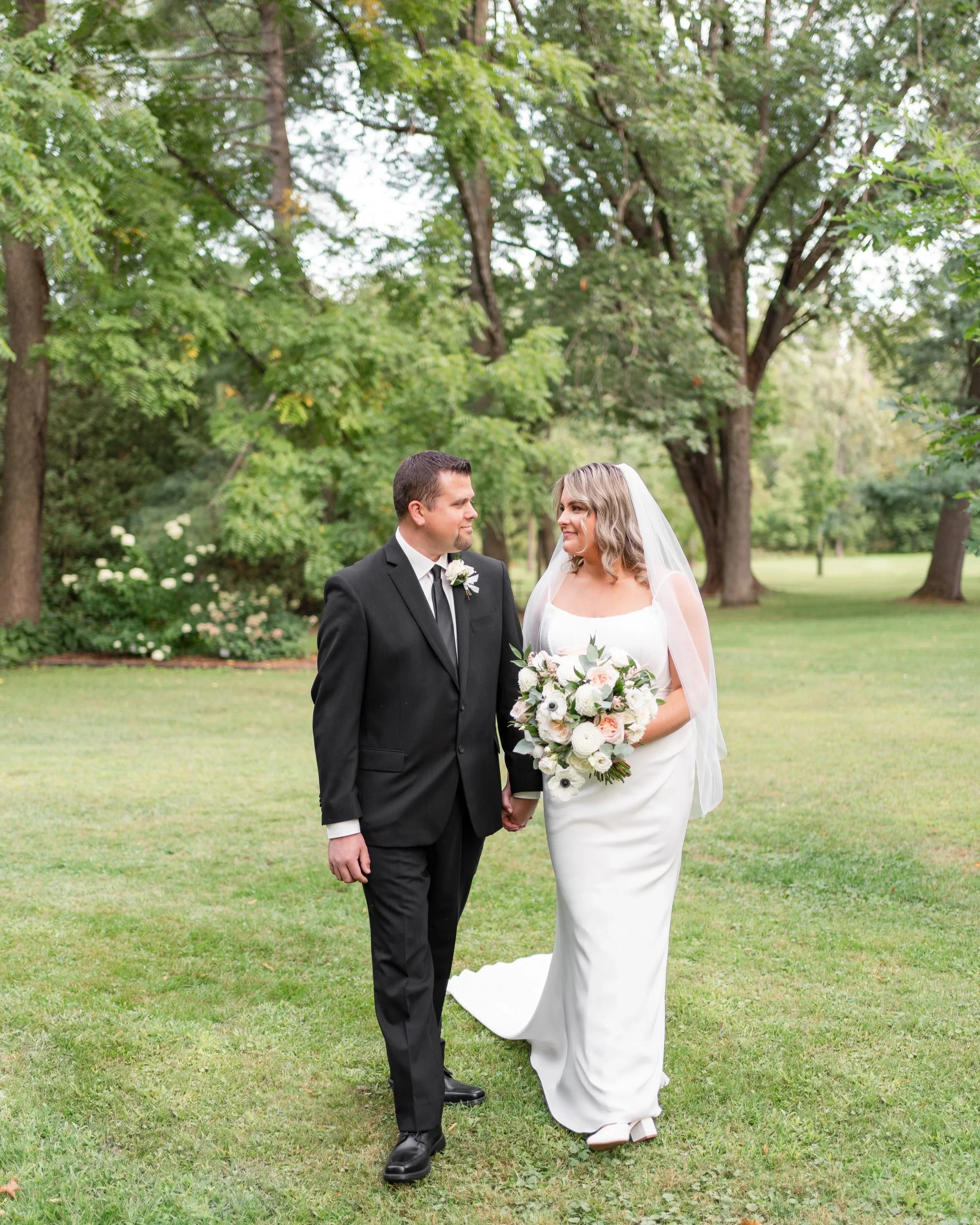A bride and groom holding hands and looking at each other outside in a park with green trees and grass. Pembroke, Ontario.