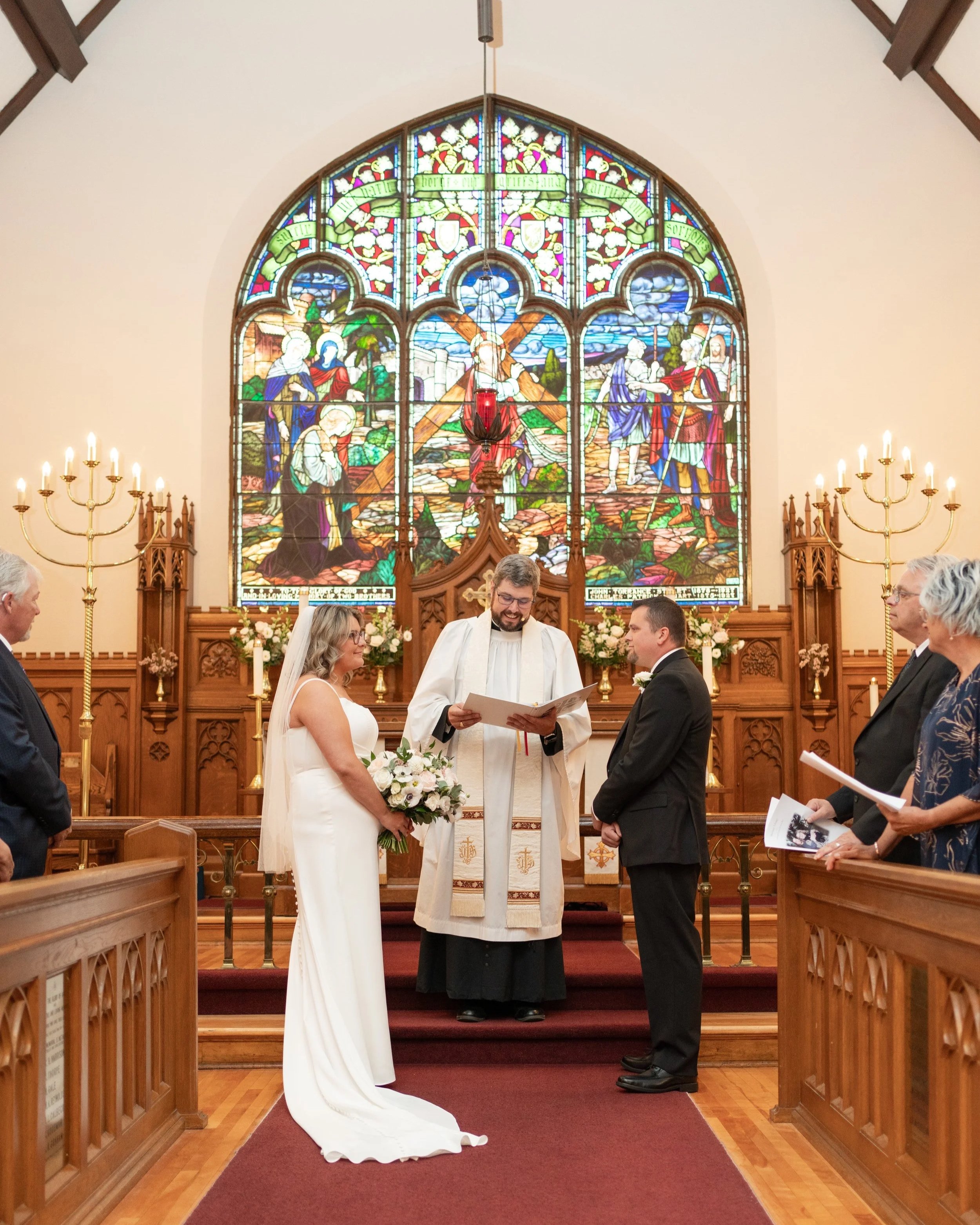 A wedding ceremony taking place in a churchThe bride and groom stand before a priest. The bride is holding a bouquet of flowers, and the groom is dressed in a black suit. Pembroke, Ontario church.