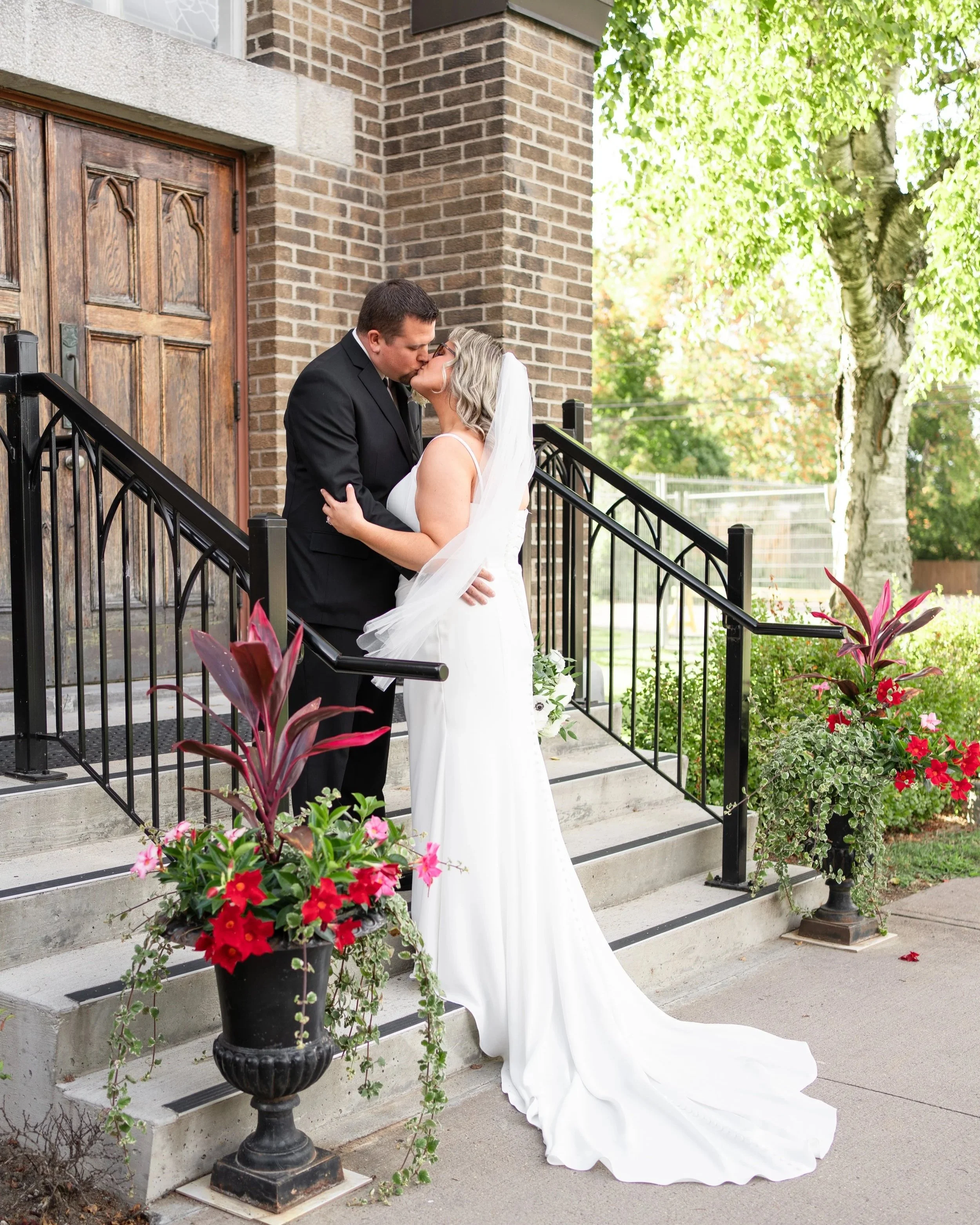 A newlywed couple sharing a kiss on the steps outside a brick building, with colorful flowers in planters nearby.