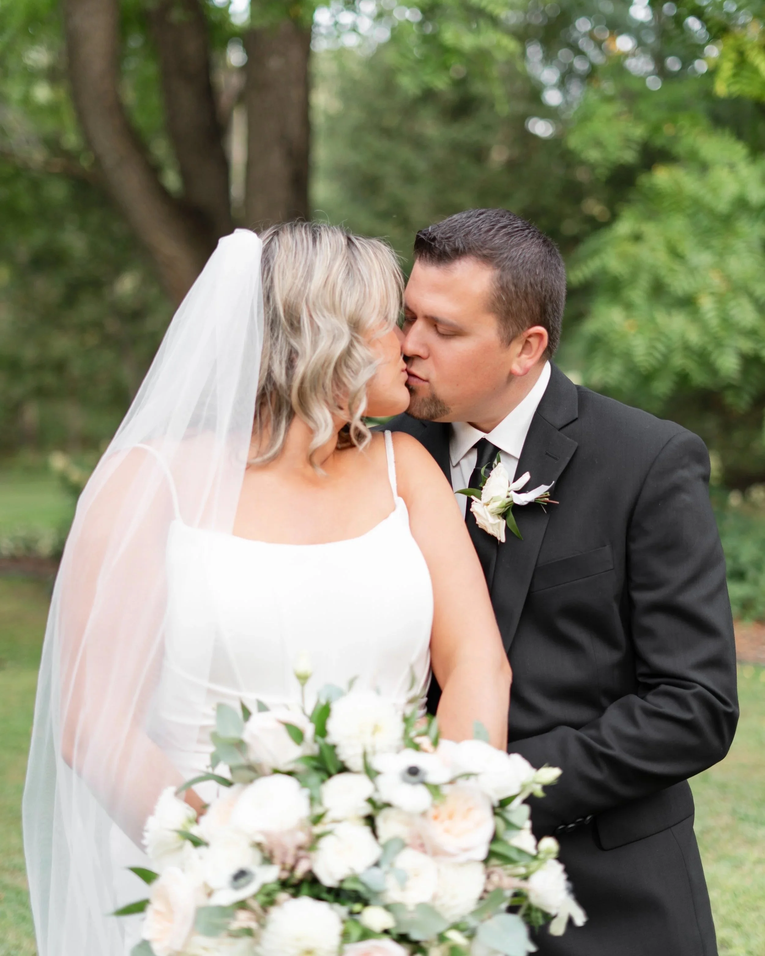 A bride and groom share a kiss outdoors, with the bride holding a large bouquet of white flowers, dressed in a white wedding gown and veil, and the groom in a black tuxedo with a white boutonniere, against a background of green trees.