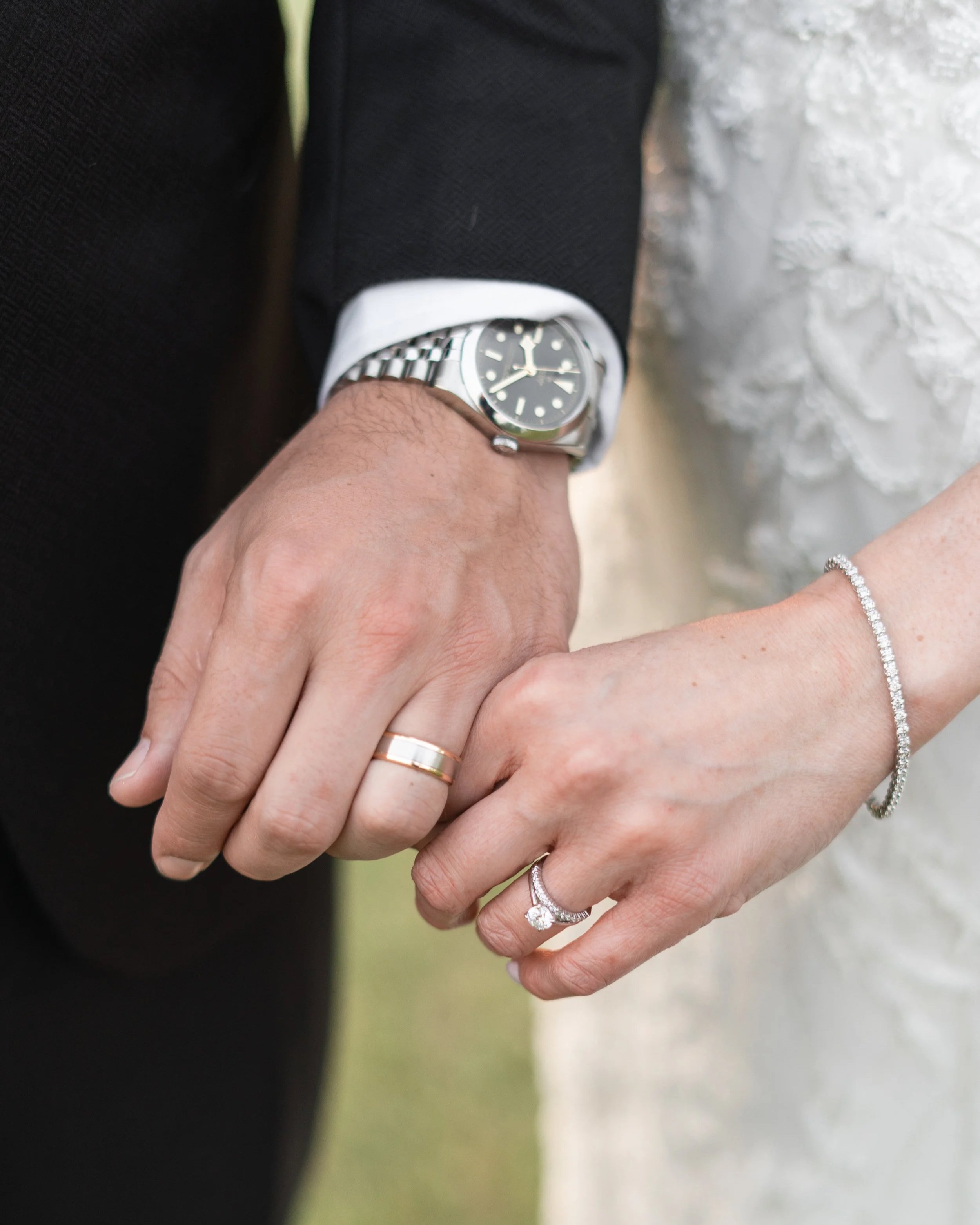Close-up of a bride and groom holding hands, showing wedding rings and jewelry. The groom wears a black suit and a wristwatch. The bride has a diamond bracelet on her wrist and a diamond engagement ring.