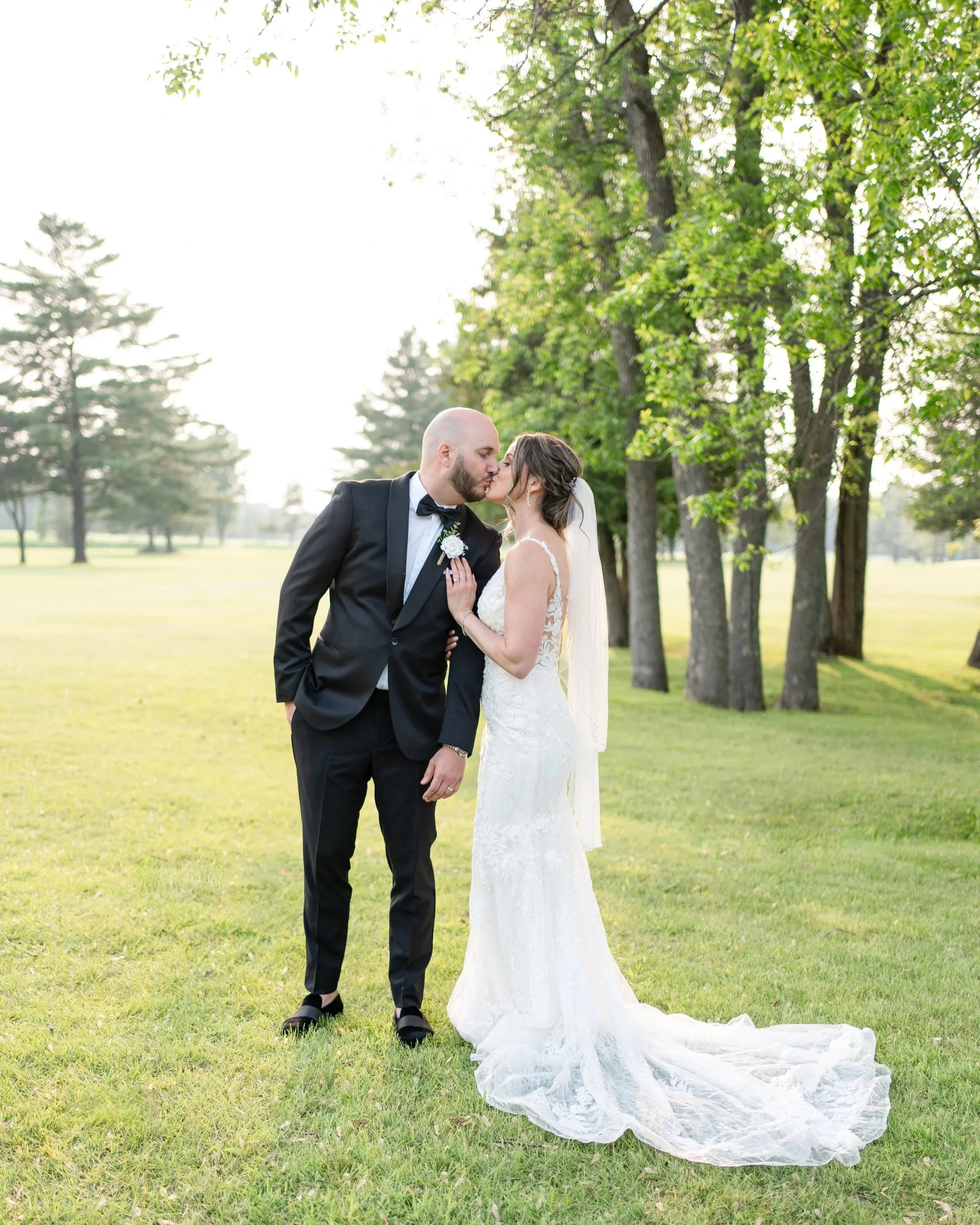 A bride and groom share a kiss on their wedding day outdoors in a park with green grass and tall trees in the background.