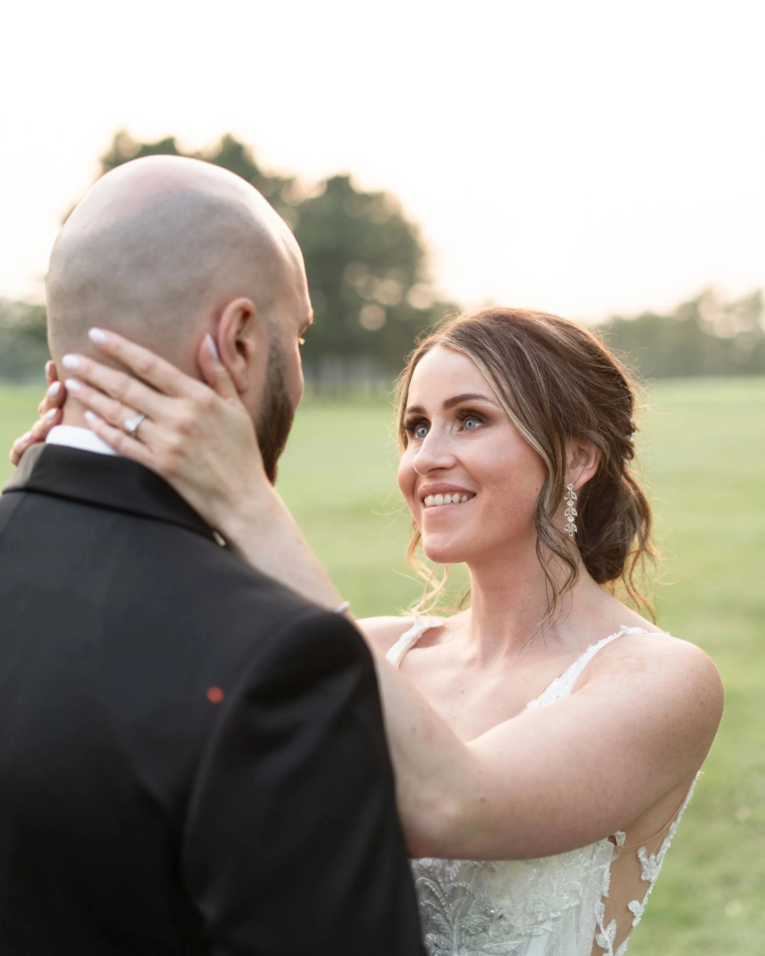 A bride and groom outdoors, embracing, with the bride smiling at the groom, on a grassy field during sunset.