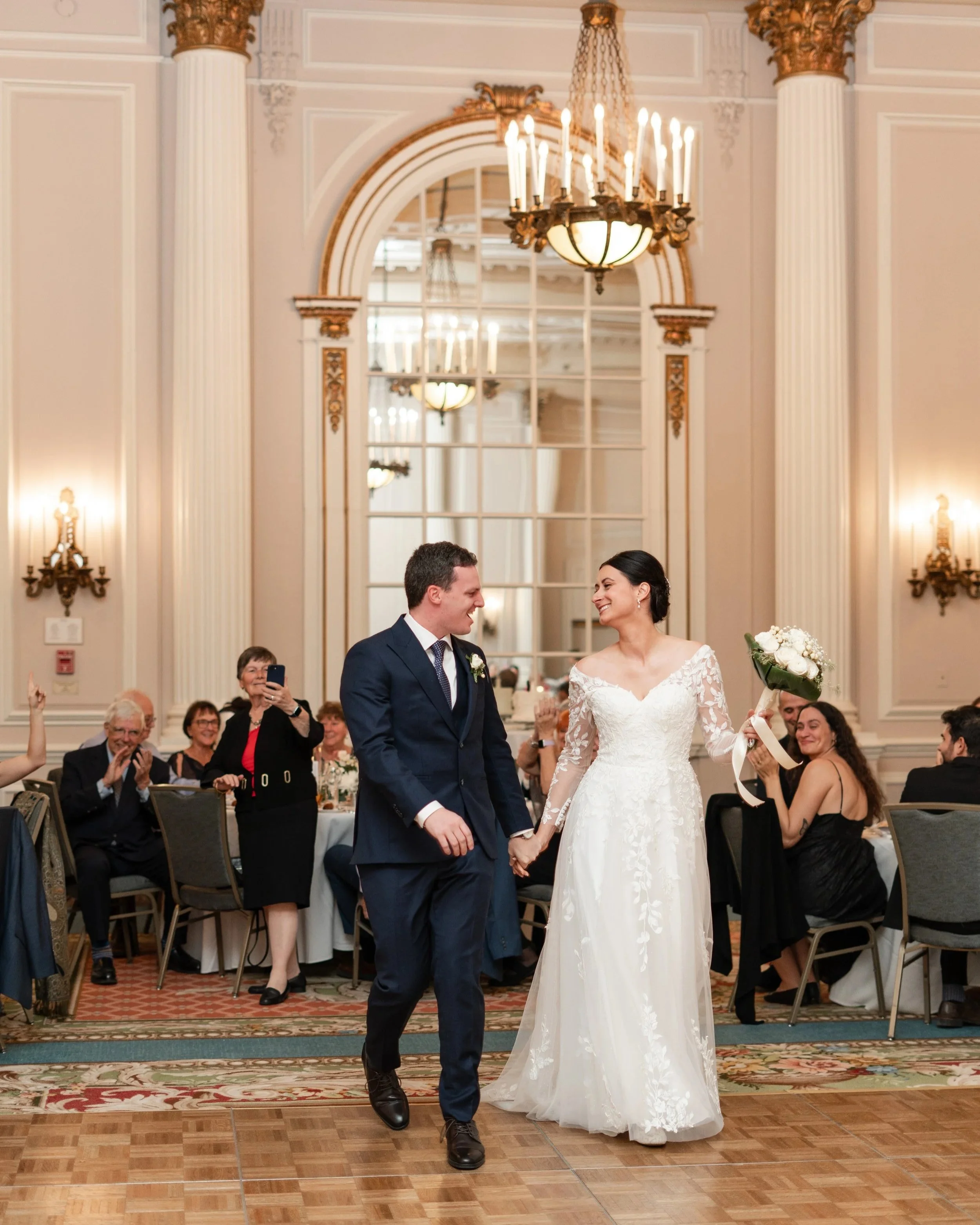 A bride and groom holding hands and smiling during their wedding reception. The bride is wearing a white lace wedding gown and holding a bouquet, while the groom is dressed in a dark blue suit. Chateau Laurier, Ottawa, ON