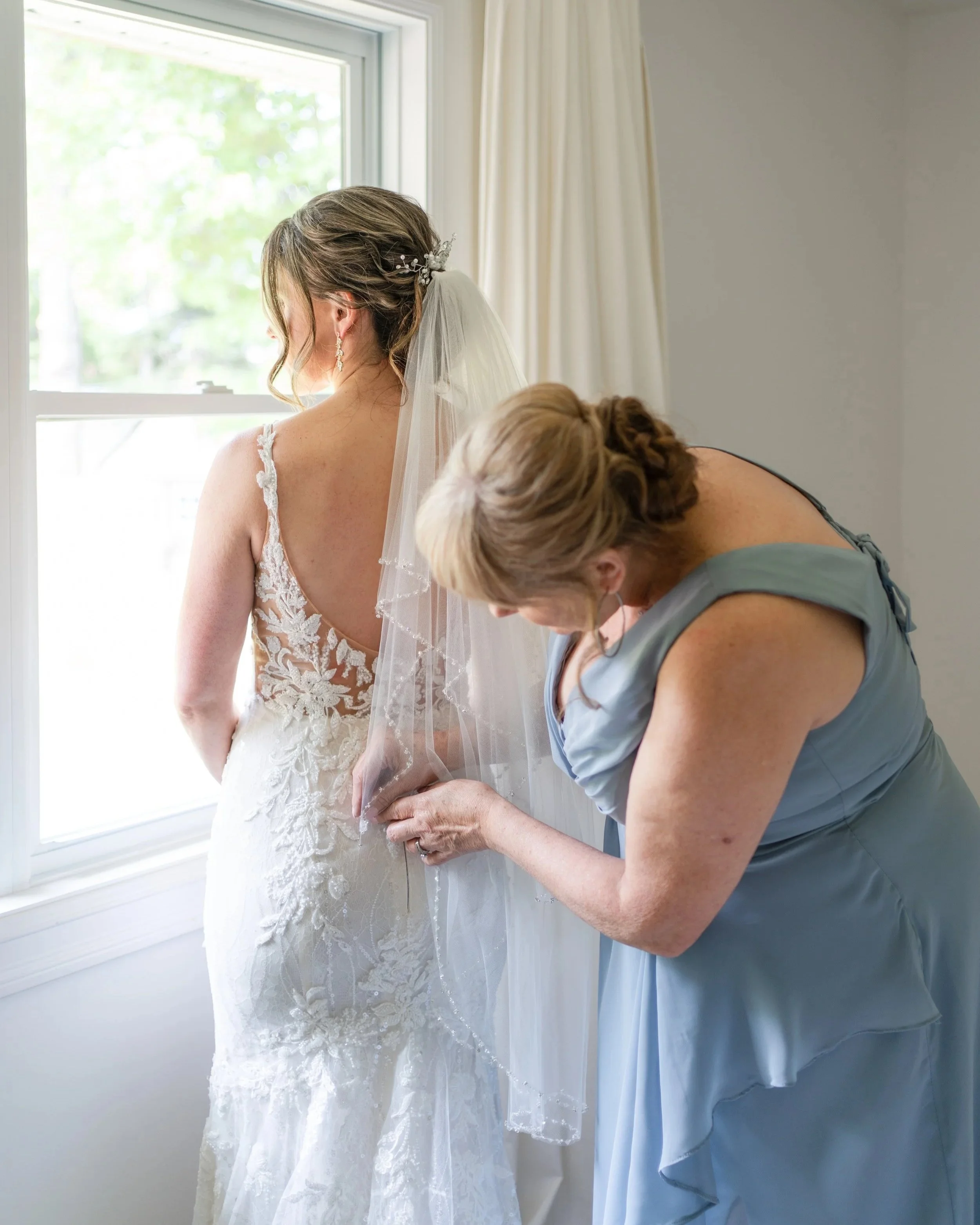 A bride getting her wedding dress adjusted by her mother in a light blue dress.