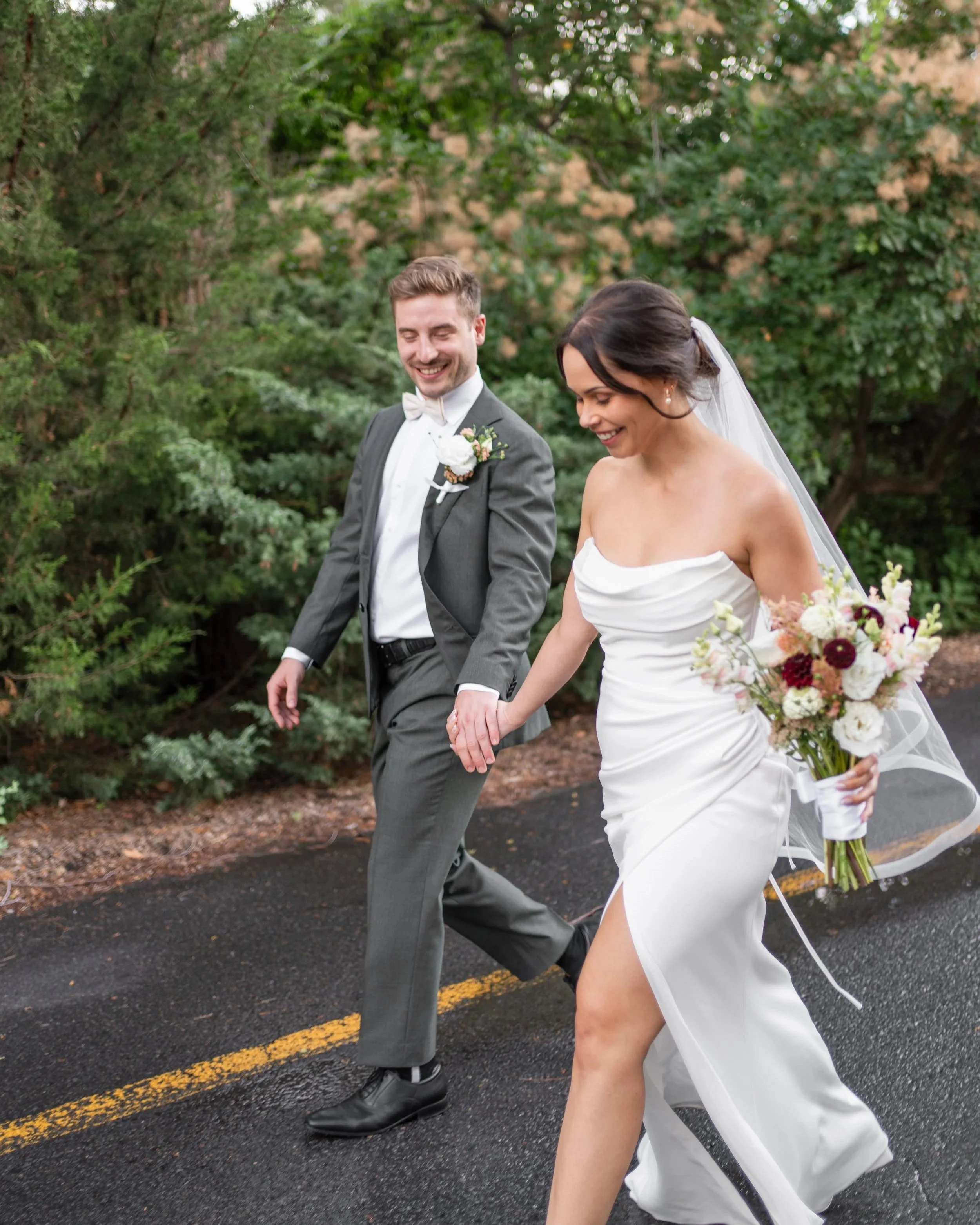 A newlywed couple holding hands and walking on a paved road, the bride in a strapless white wedding dress with a bouquet of flowers, and the groom in a gray suit with a white shirt and bow tie, both smiling and looking down. La Toundra, Montreal
