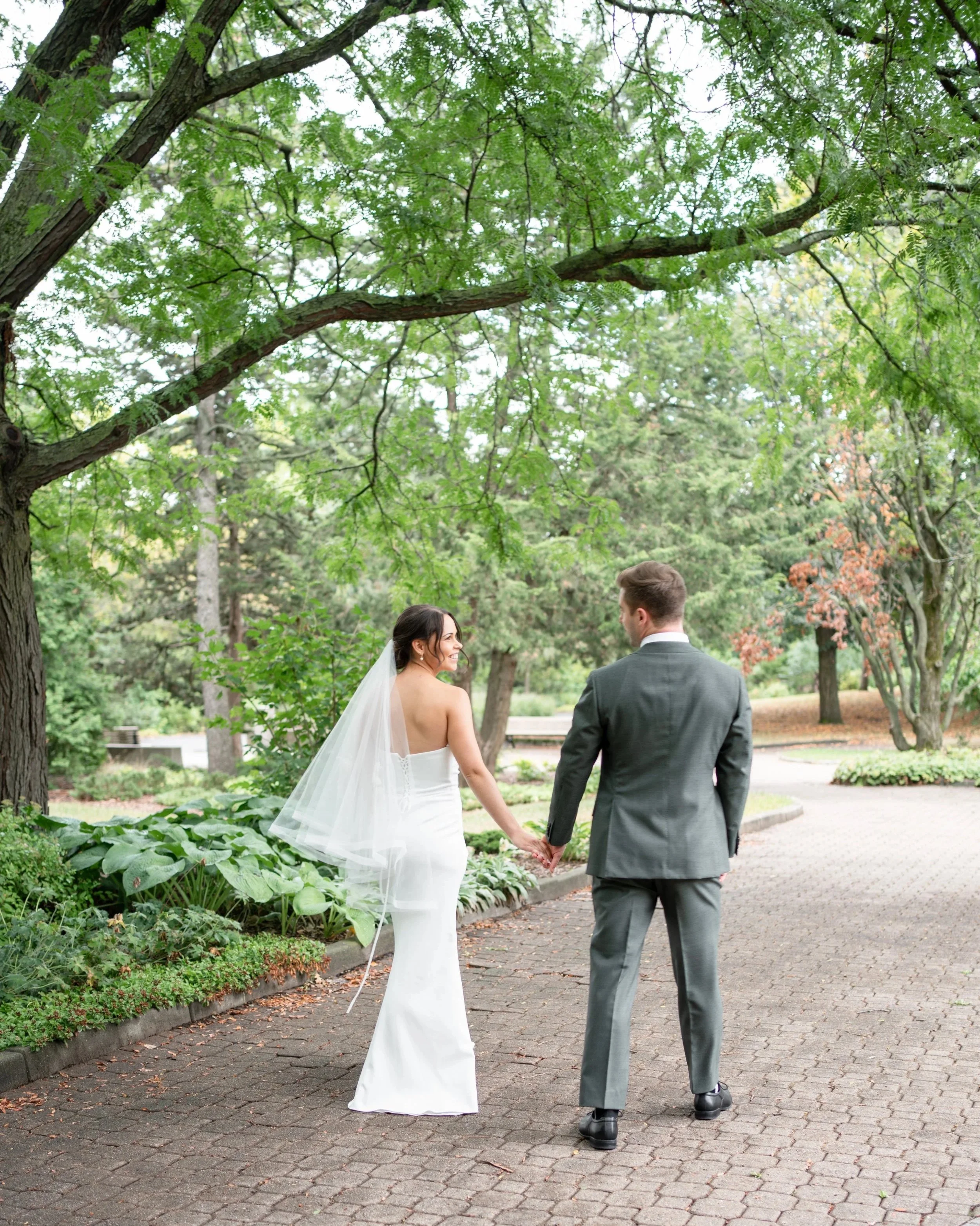 A newlywed couple holding hands and walking in a lush park with green trees and plants, the bride wearing a white wedding dress and veil, the groom in a gray suit, both smiling. La Toundra, Montreal