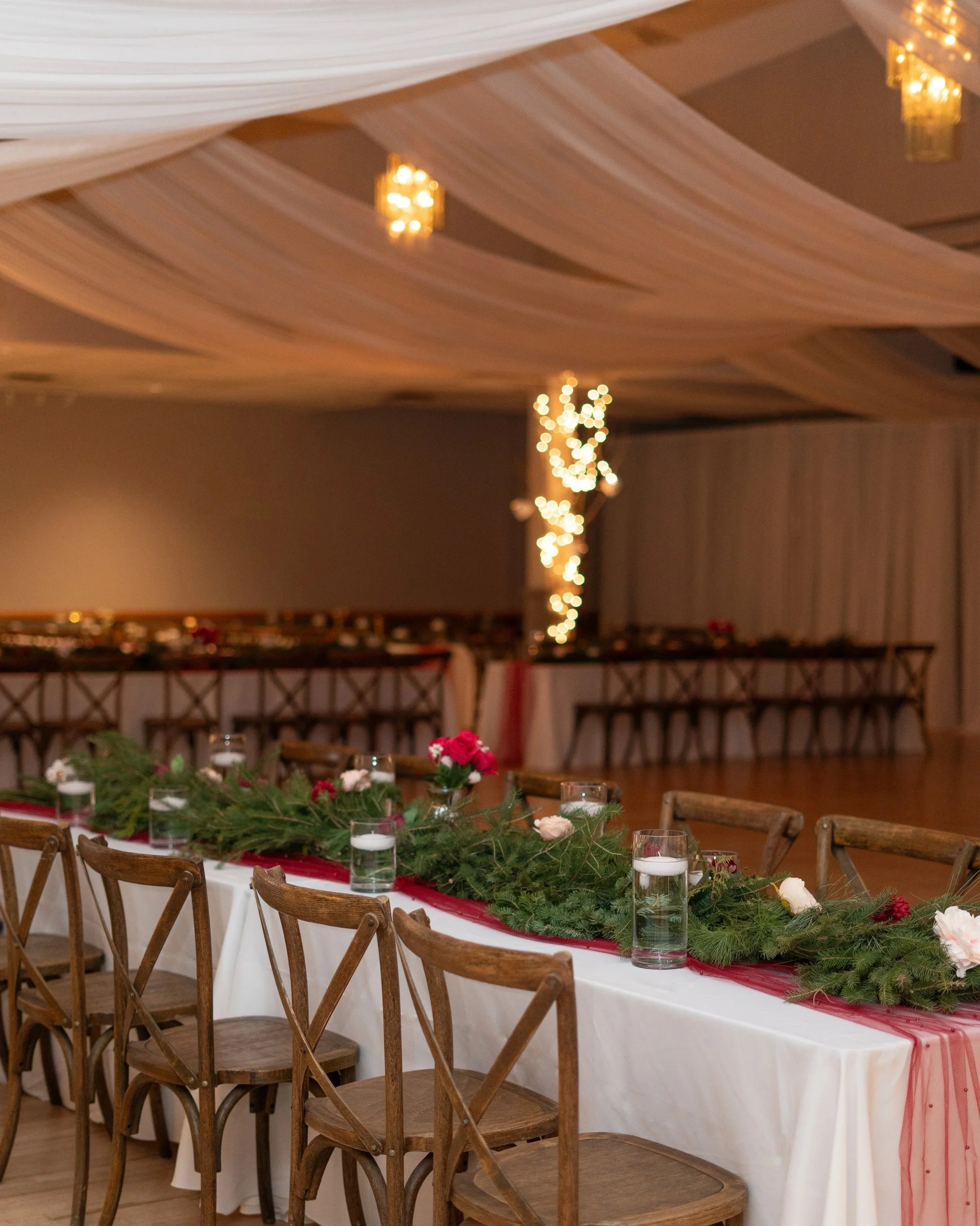 Decorated banquet table with greenery, pink and white flowers, candles, and a white tablecloth, set in a decorated hall with draped ceiling fabric and warm hanging lights. Pemrboke Germanian Hall