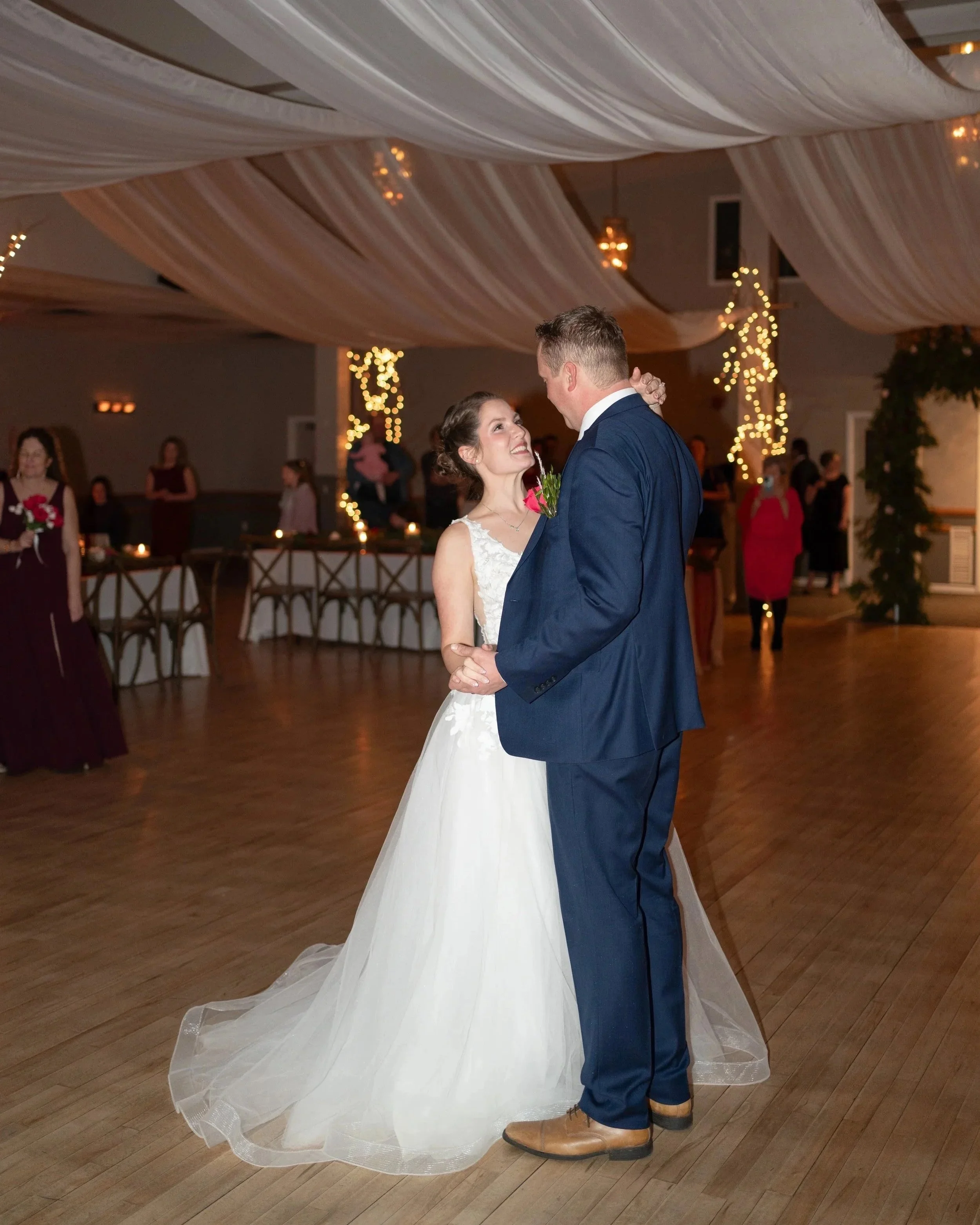 A bride and groom sharing their first dance at a wedding reception, surrounded by guests and decorated with fairy lights and draped fabric ceiling.