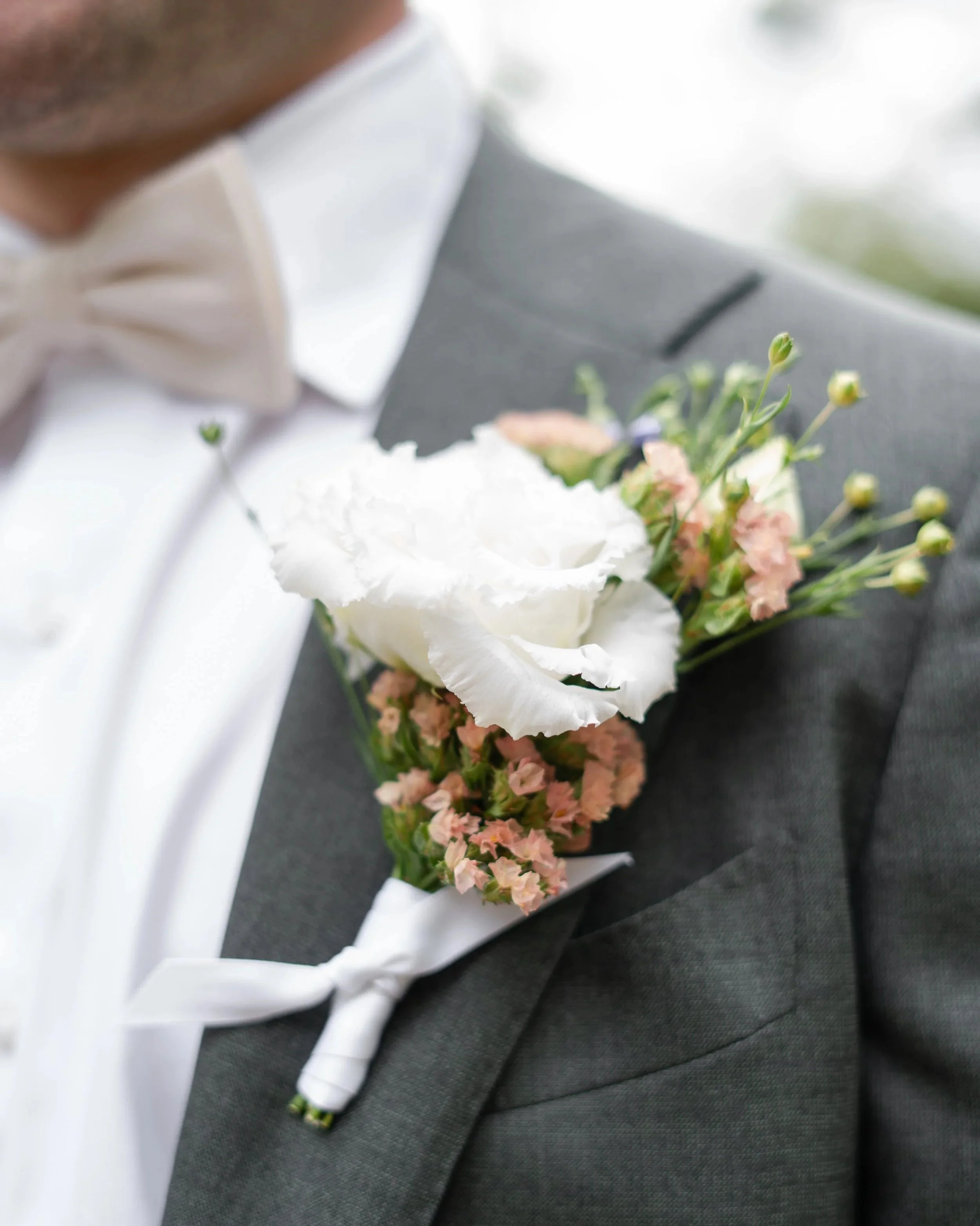 Close-up of a man's suit with a boutonniere pinned to his lapel, consisting of a white flower and small pink flowers tied with a white ribbon.