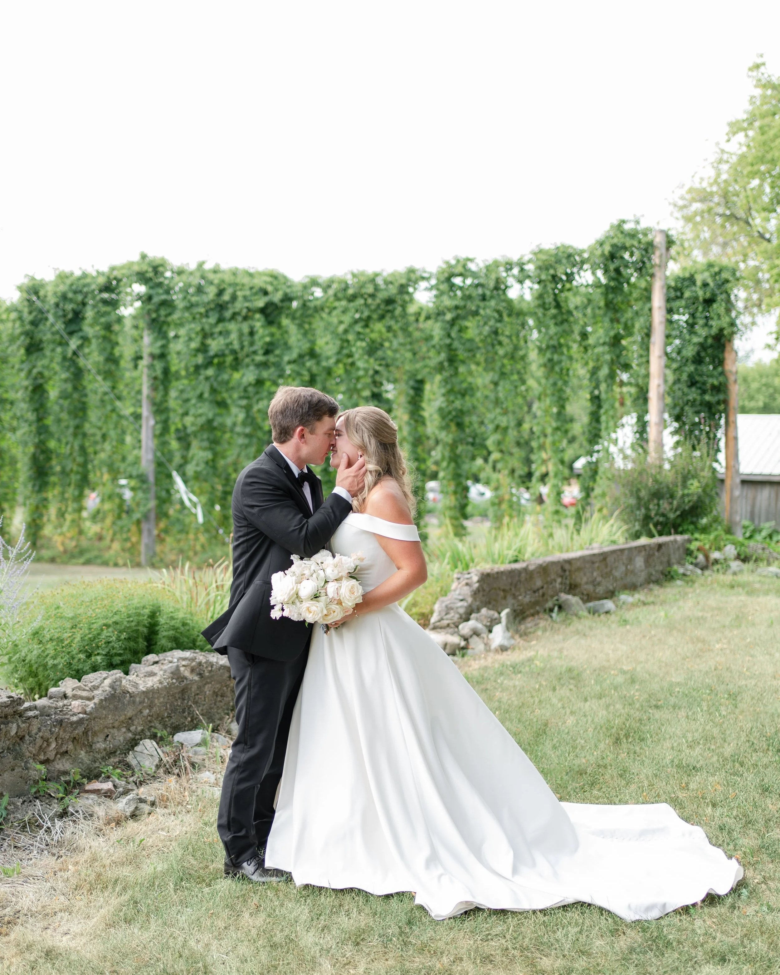 A bride and groom share a romantic moment, touching foreheads outdoors in a garden setting. Stone Hedges Hops, Pembroke, ON
