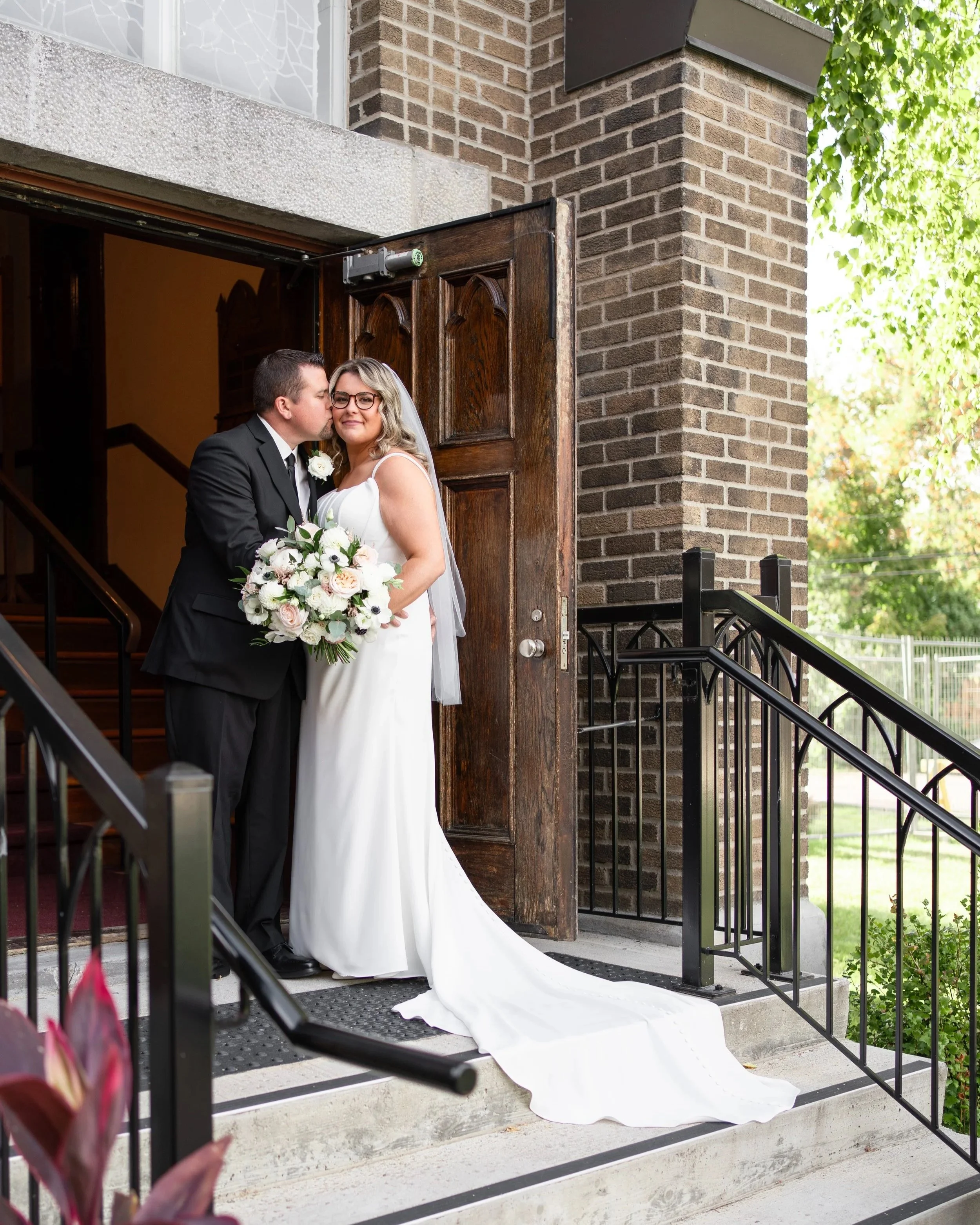 A bride and groom standing in the doorway of a church, with the groom kissing the bride on the cheek. The bride is holding a bouquet of white and light pink flowers, and her wedding dress has a long train. Pemrboke church