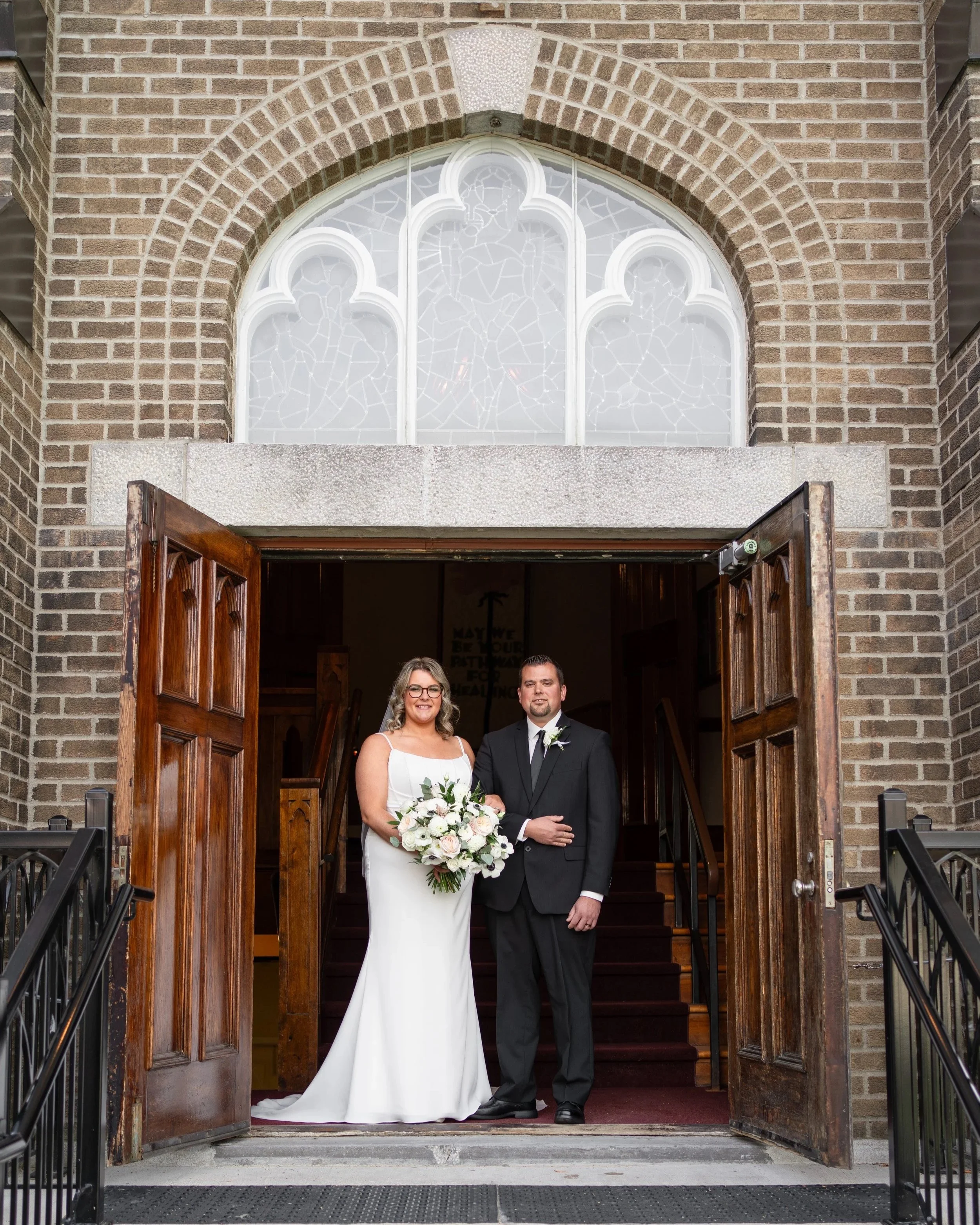 A bride and groom standing at the entrance of a church, with open wooden doors behind them. The bride is wearing a white wedding dress and holding a bouquet of flowers, and the groom is in a black suit with a white shirt and black tie. The church has