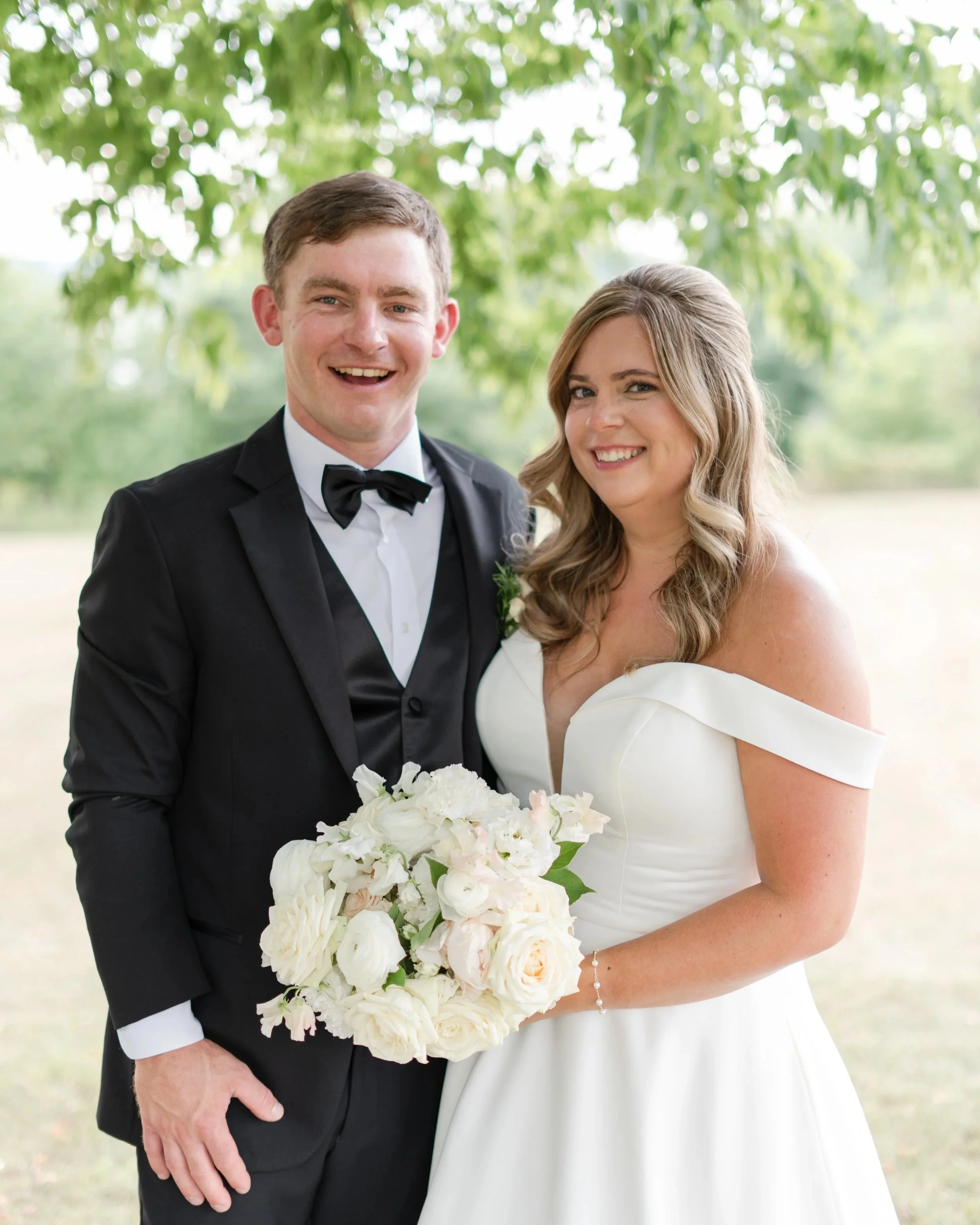 A happy bride and groom stand outdoors on their wedding day, with the bride holding a bouquet of white flowers and wearing an off-shoulder white wedding dress, and the groom dressed in a black tuxedo with a bow tie, smiling at the camera.