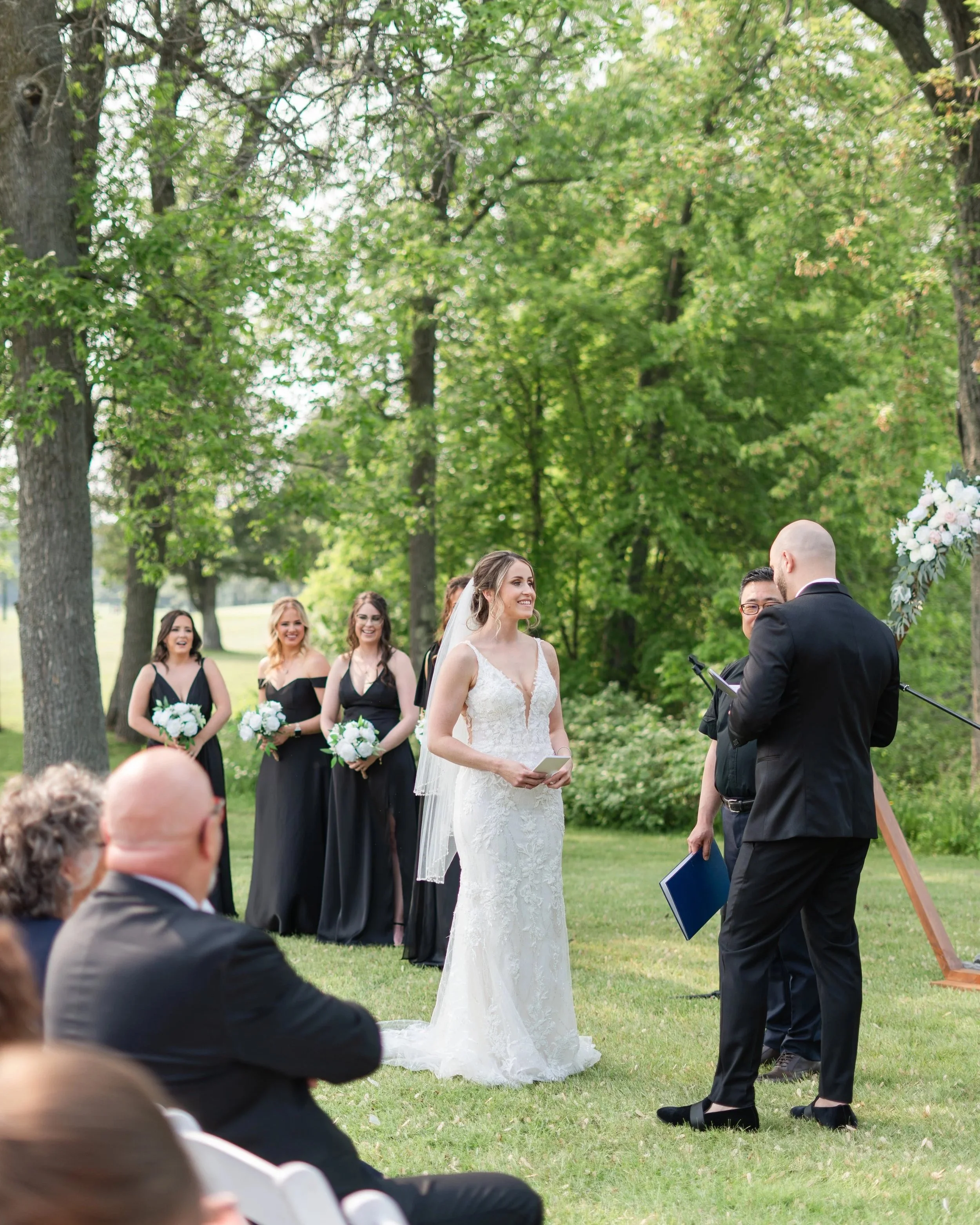 A bride in a white lace wedding dress is standing outdoors during a wedding ceremony, smiling at the officiant. Behind her, three bridesmaids in black dresses hold bouquets of white flowers, smiling. Guests are seated and watching the ceremony in a l