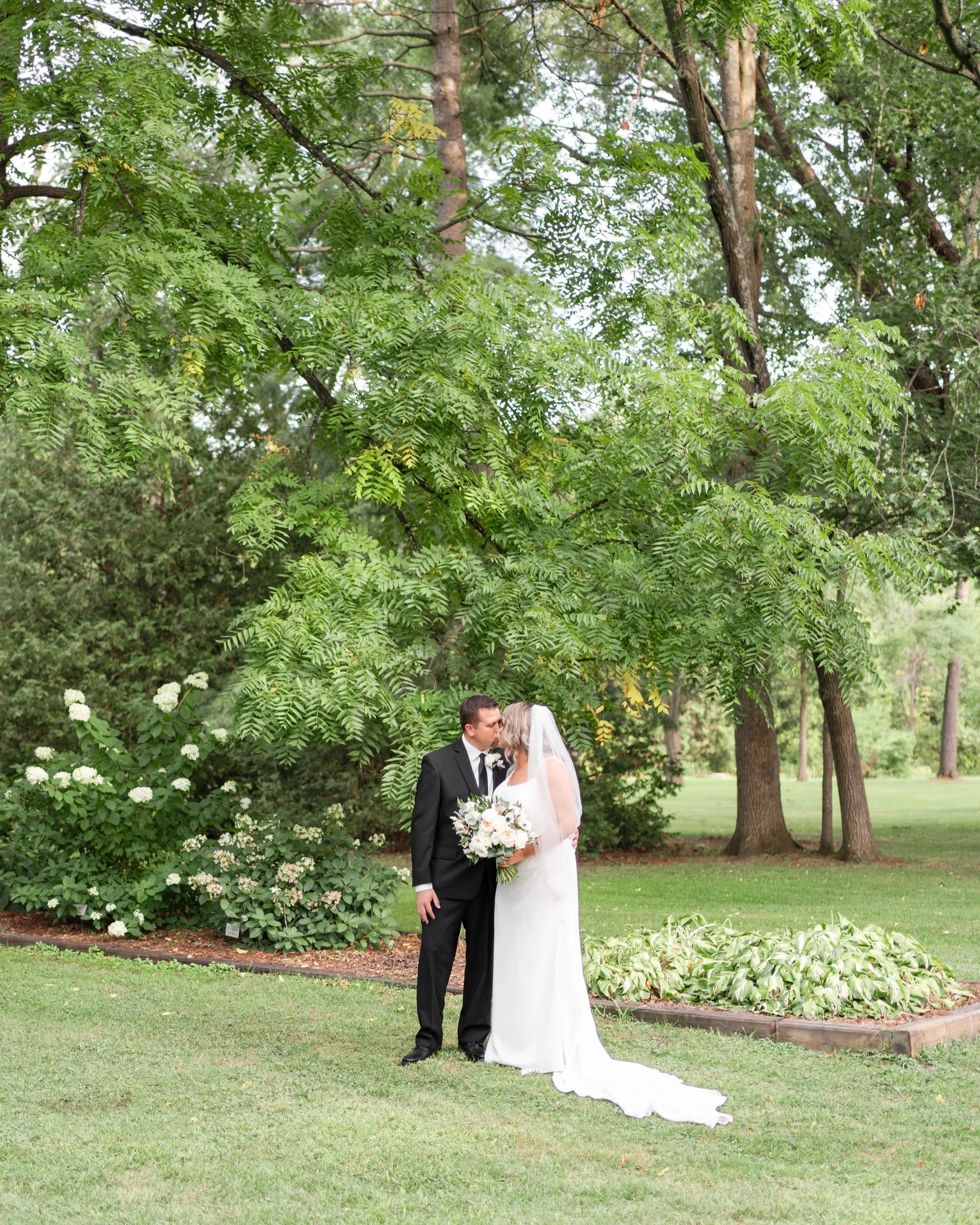 A bride and groom share a kiss outdoors in a park setting, surrounded by green trees and bushes, with a flower bed nearby. Wedding in Pembroke, Ontario