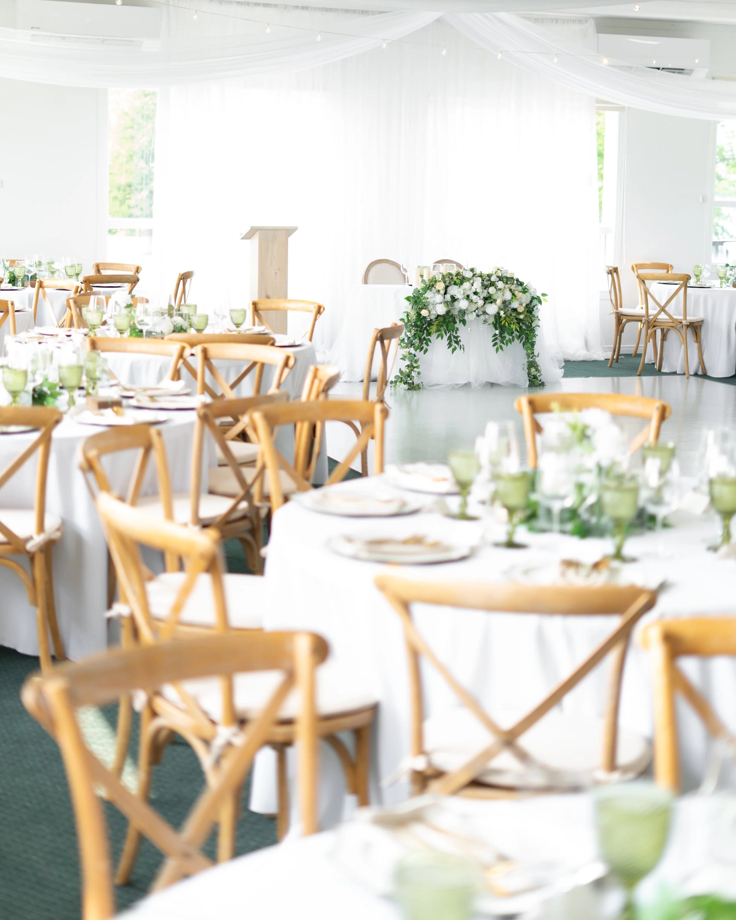 Wedding reception hall with round tables covered in white tablecloths, set with plates, utensils, glasses, and green cups. Wooden chairs surround the tables. The room has white drapes and light coming through windows. Pembroke Golf Club