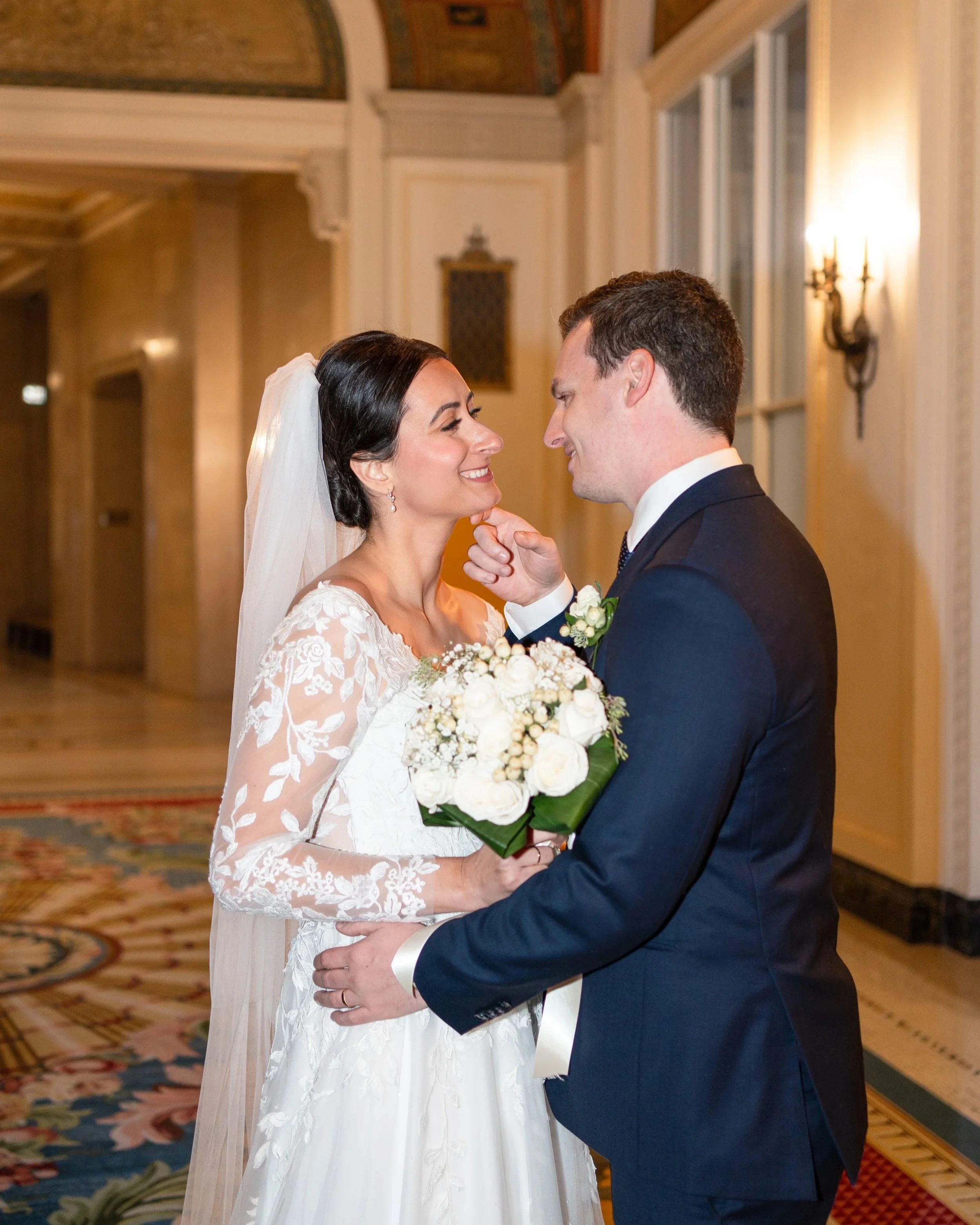 A bride and groom on their wedding day in a decorated indoor venue, sharing a joyful moment as the groom touches the bride's chin. The bride wears a white lace wedding dress and veil, holding a bouquet of white flowers. The groom is in a dark suit wi