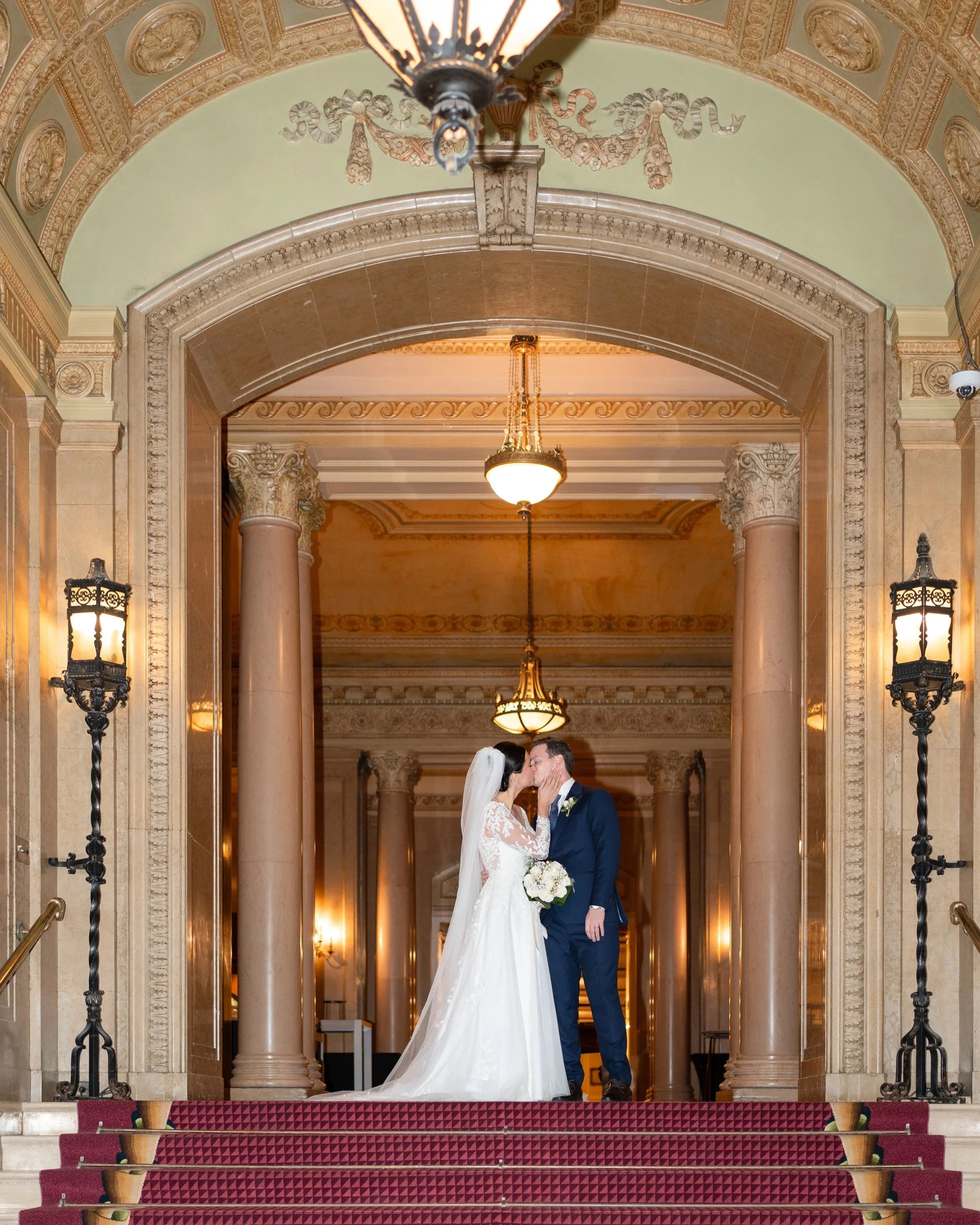 A bride and groom sharing a kiss on the staircase of a grand, ornate building with classical architecture and elegant lighting.  Wedding at Chateau Laurier in Ottawa, Ontario