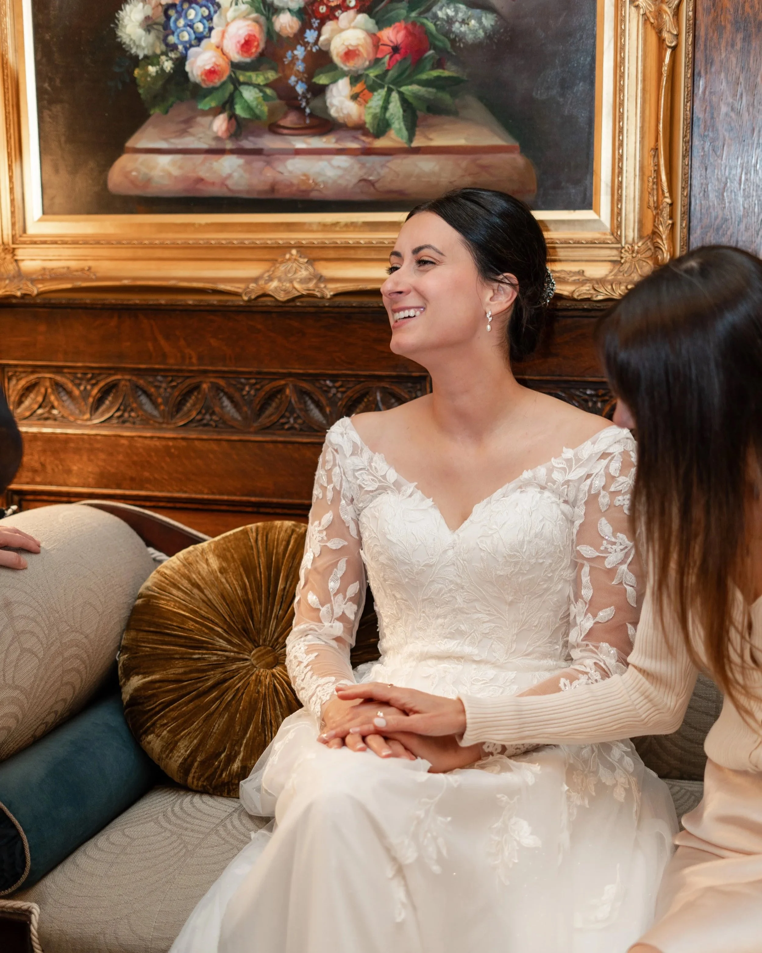 A bride with dark hair in an elegant white lace wedding dress, sitting on a vintage sofa with a gold-framed floral painting in the background. Wedding at Chateau Laurier in Ottawa, Ontario
