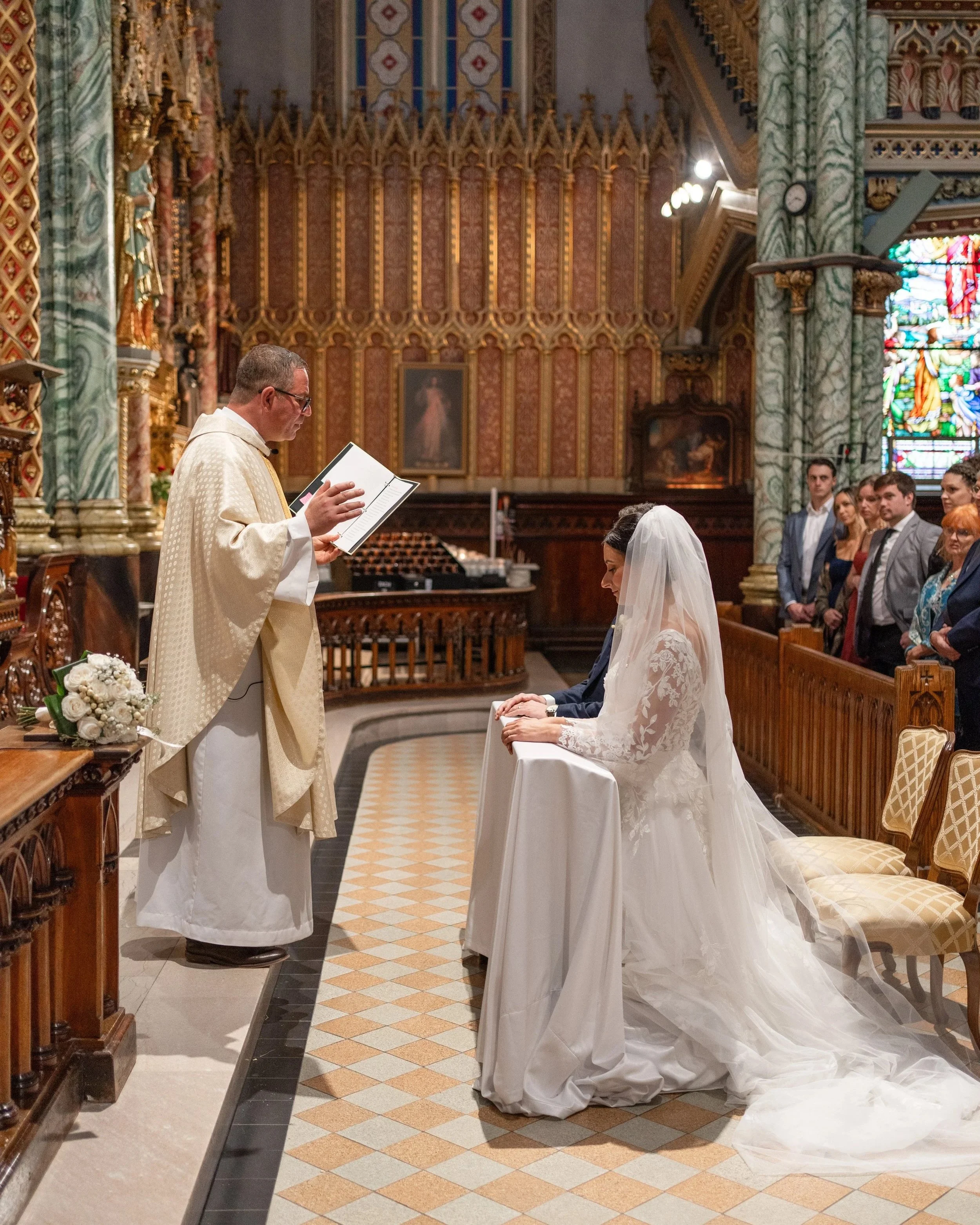 A wedding ceremony inside a church with ornate gold and red decor. The bride in a white lace wedding gown and veil is kneeling at a small table, praying, while the priest reads from a book. In the background, guests are seated and watching the ceremo