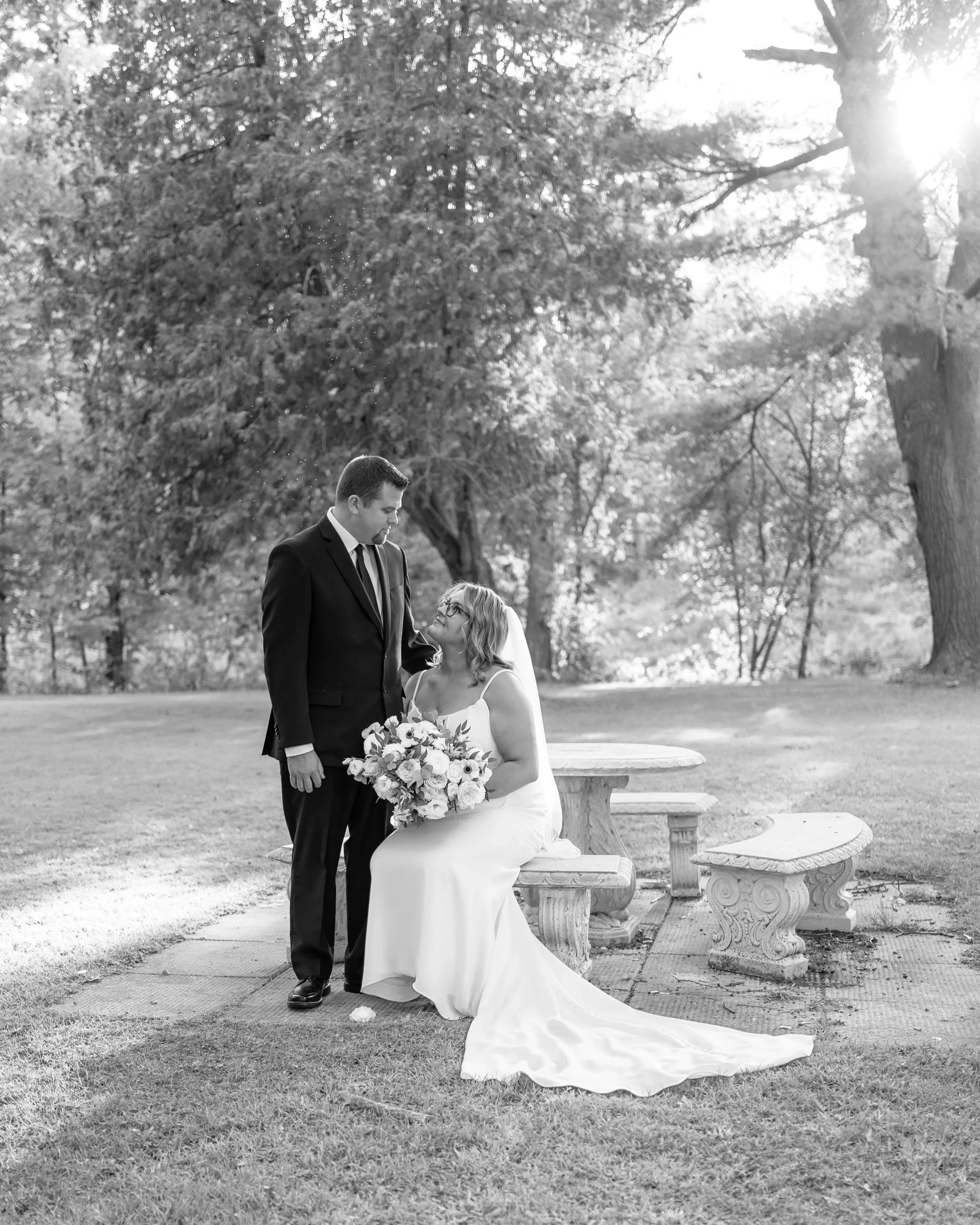 A black and white photo of a bride and groom outdoors, with the bride sitting on a stone bench holding a bouquet of flowers, and the groom standing next to her, looking at her, with trees and sunlight in the background. Wedding in Pembroke, Ontario