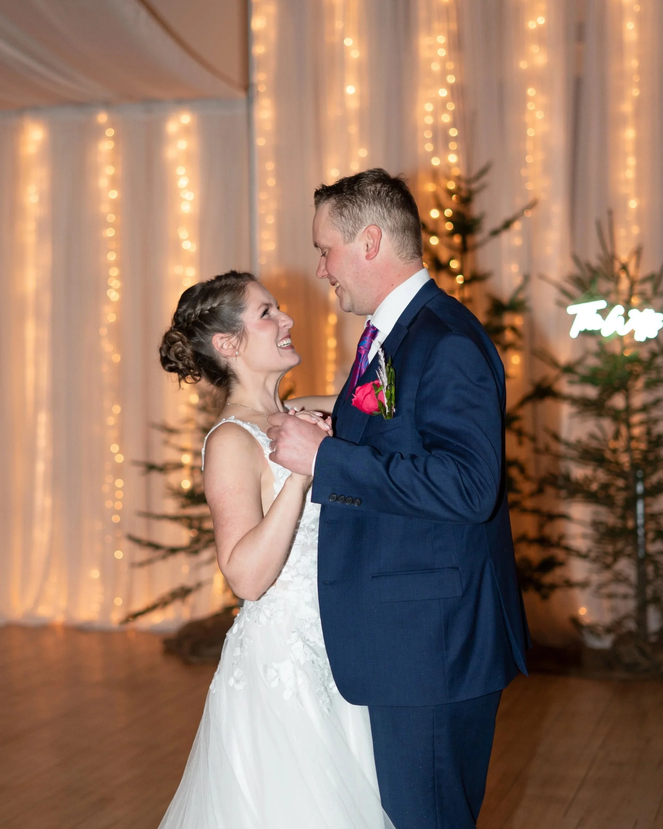 A bride and groom share a dance at their wedding reception, smiling at each other. The bride wears a white gown, and the groom wears a navy suit with a pink boutonniere. The background features warm string lights and decorated Christmas trees. Weddin