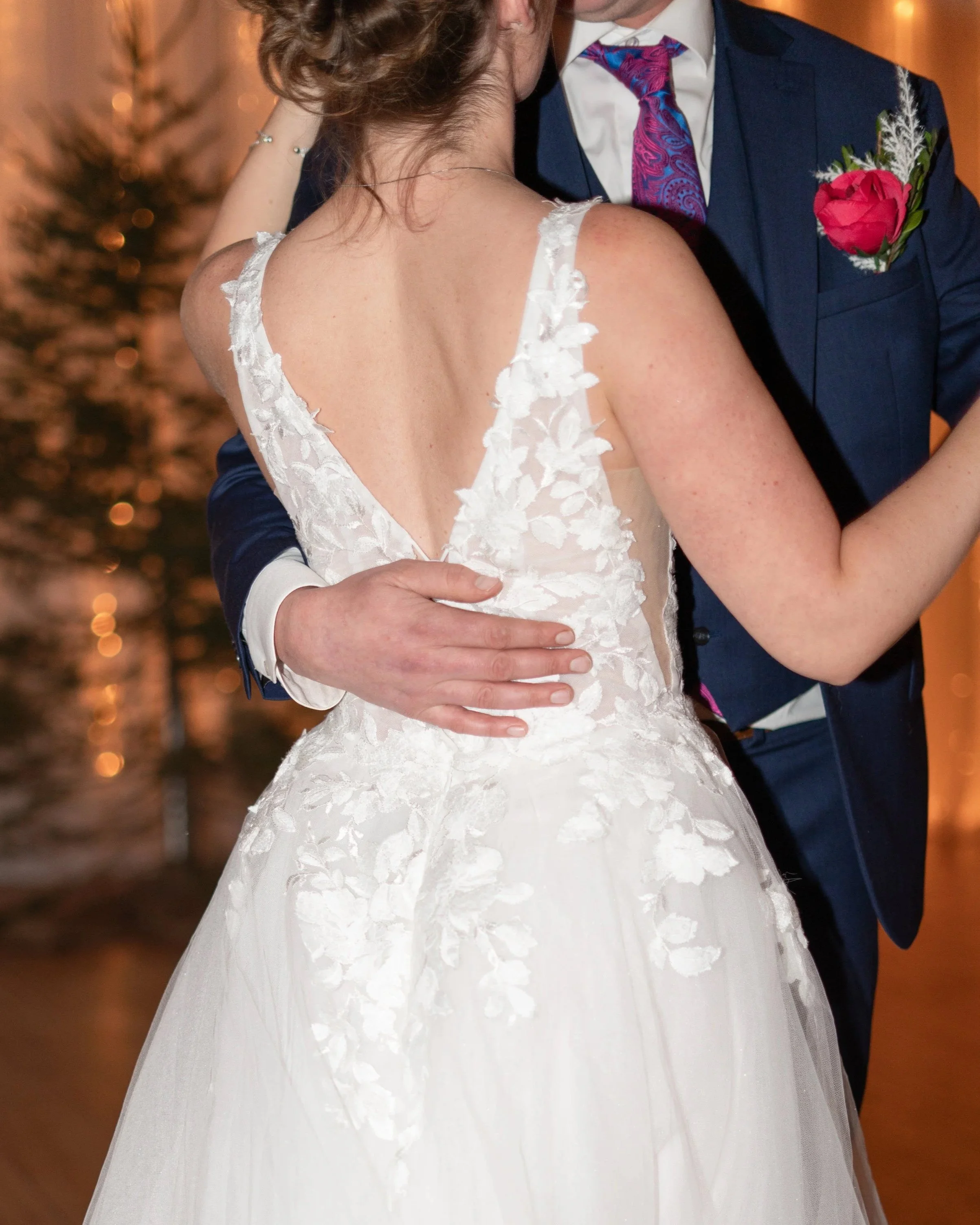 A bride in a white wedding dress dancing with a groom in a dark blue suit and pink tie, with trees and warm lighting in the background. Wedding in Pembroke, Ontario